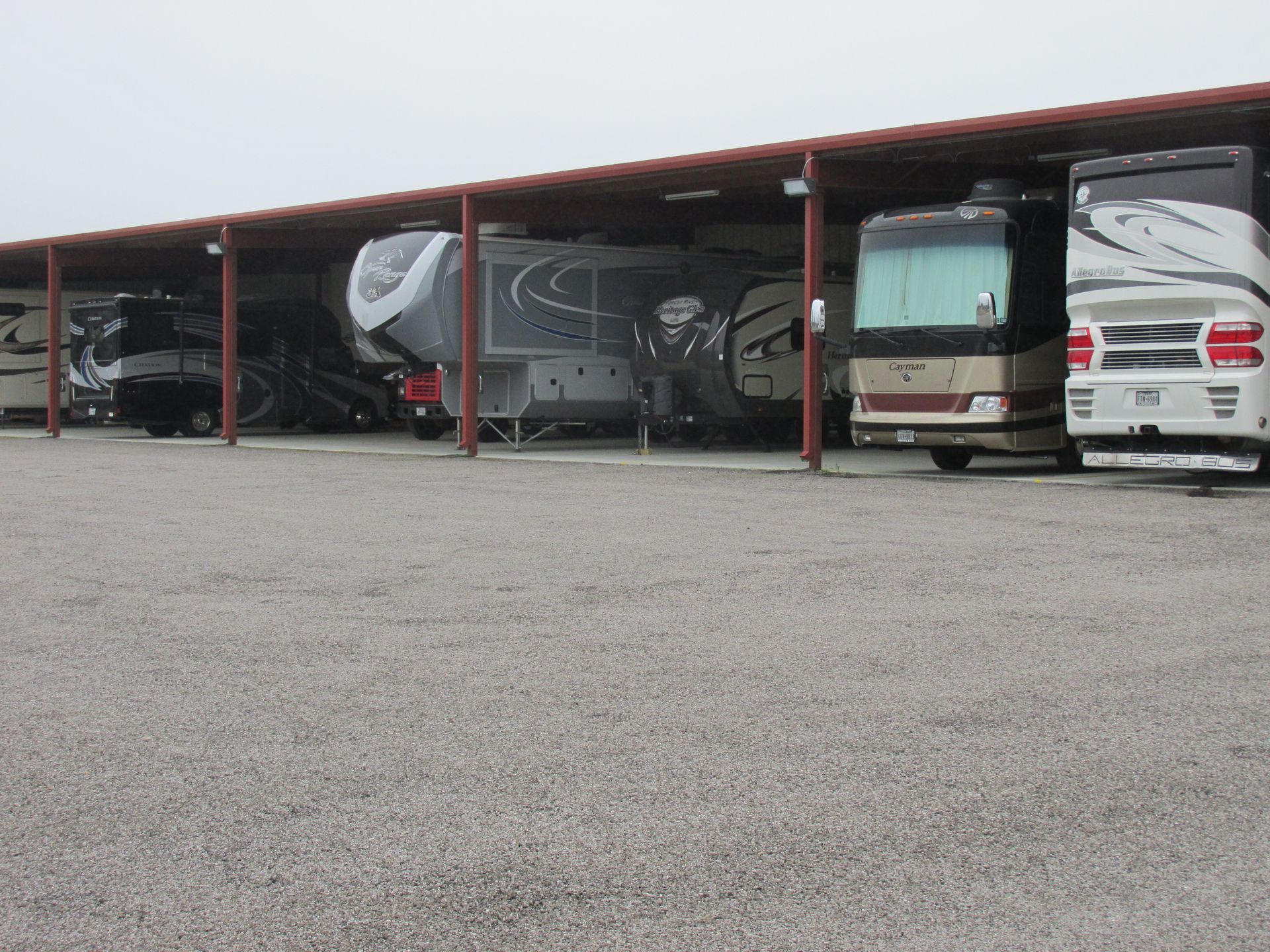 Several large RVs parked under a long, metal-roofed outdoor storage structure on a gravel lot.