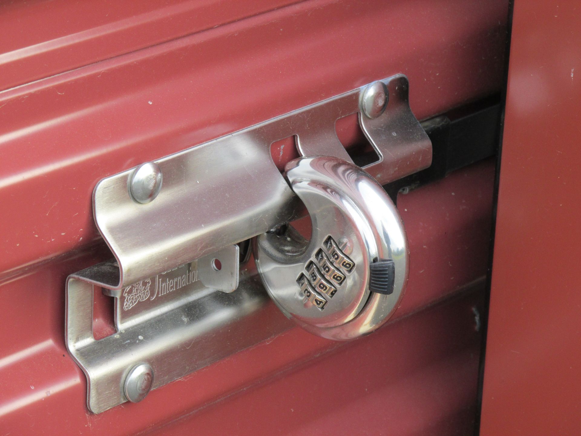 A silver disc-style combination padlock secured on the metal latch of a red storage unit door.