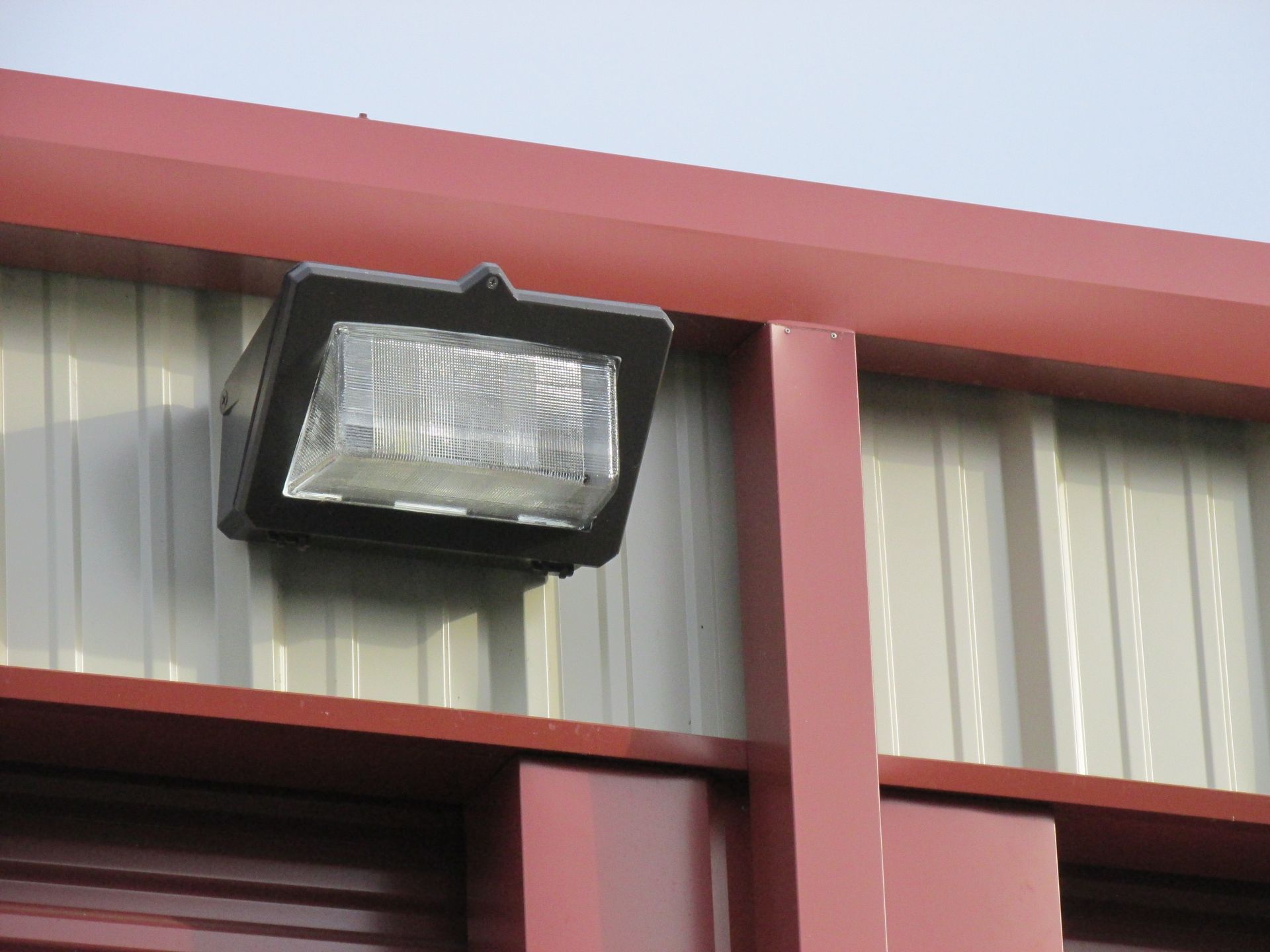 A dark-colored outdoor security light mounted on the light-colored metal siding of a building with red trim.