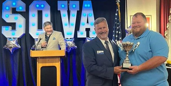 Three men at a SOVA event. One gives a trophy to a smiling man; another speaks at a podium.