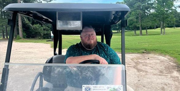 Man with a beard driving a golf cart on a grassy path. Blue shirt. Outdoor setting.