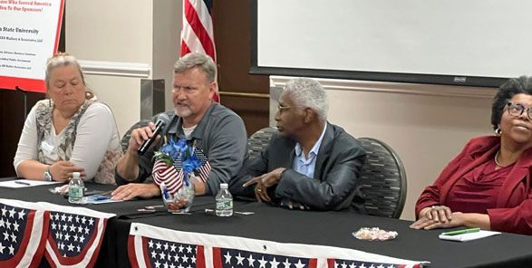 Panel of four people speaking at a table with an American flag backdrop.