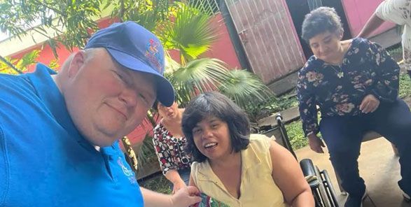 A group of people outdoors, a man in a blue shirt and hat takes a selfie with smiling women.