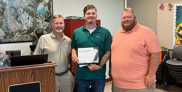Three men stand indoors; one in green shirt holds a certificate, flanked by men in tan and peach shirts.
