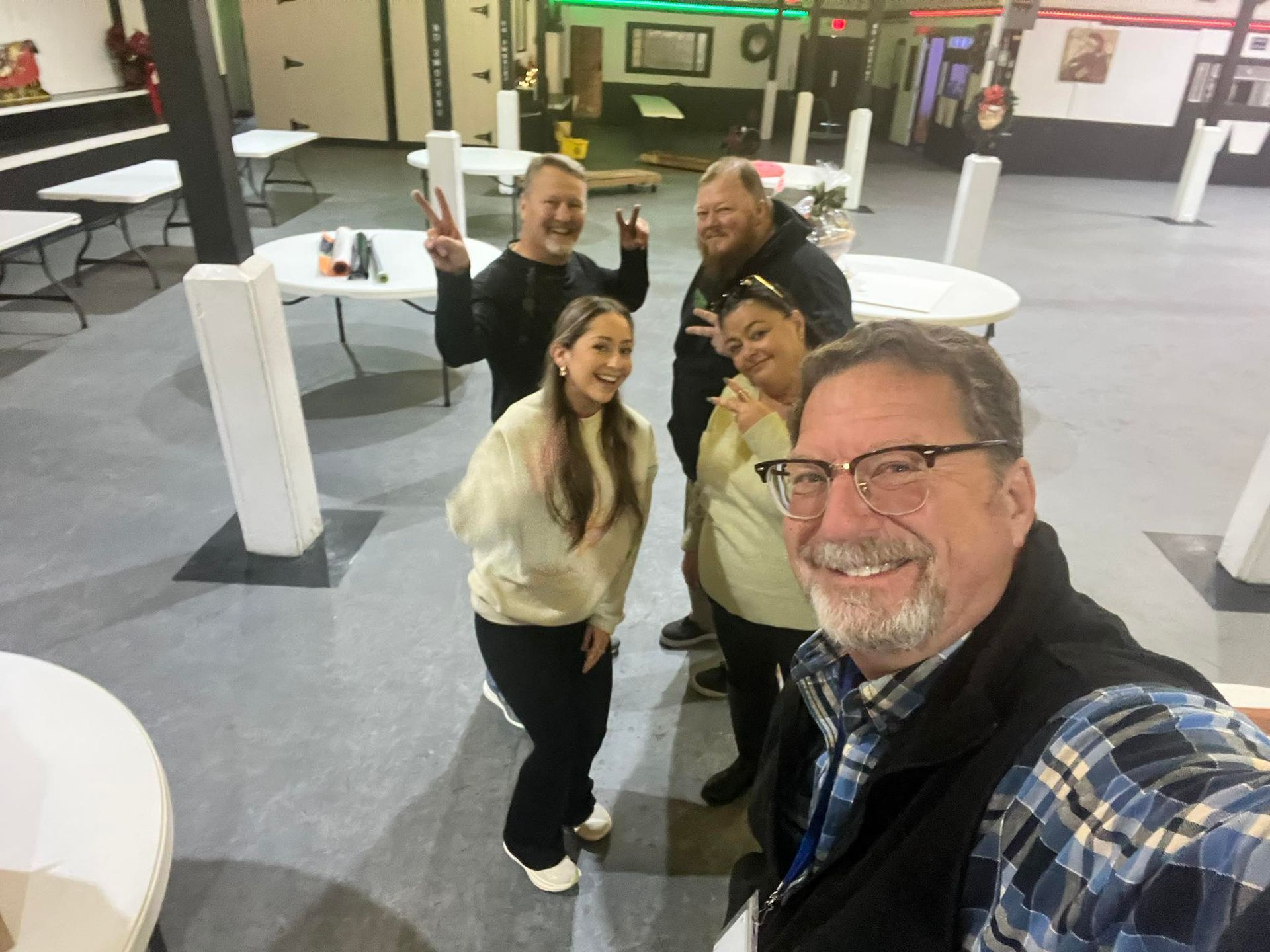 Five people pose for a selfie in an event space, smiling. Some give peace signs. White tables and posts.