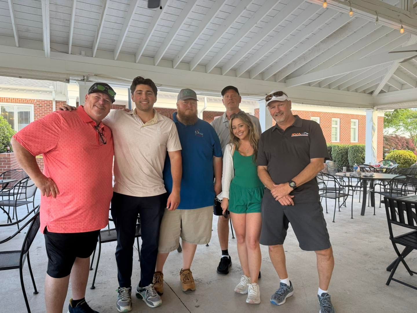 Five people posing together under a covered outdoor patio with tables and chairs in the background.