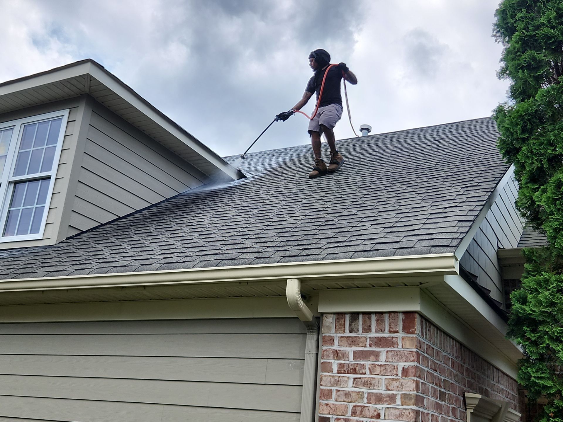 Person pressure washing a dark, textured roof on a house with beige siding and a brick section.