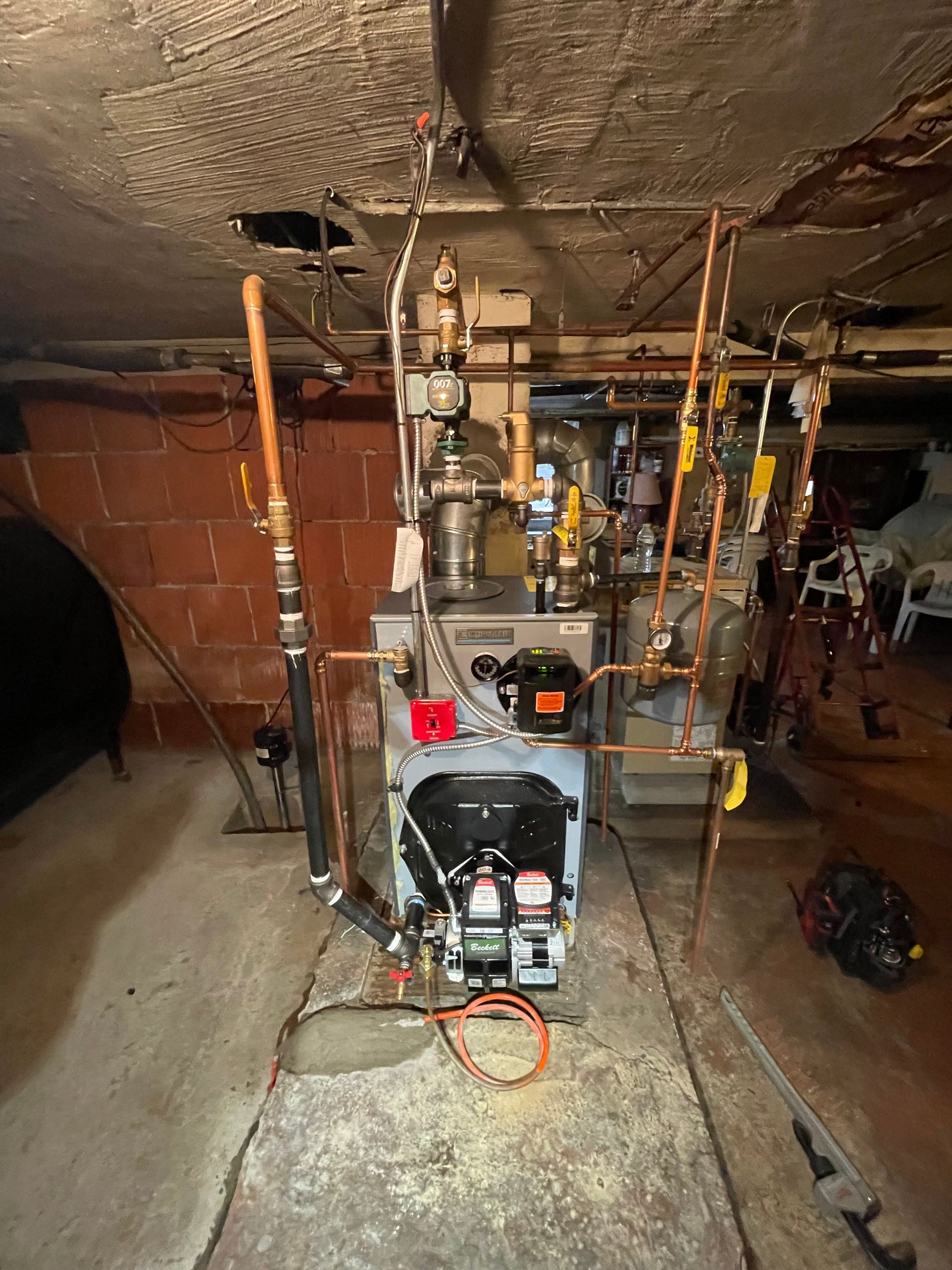 Oil-fired furnace and connected pipes in a basement setting with red brick walls and concrete floor.