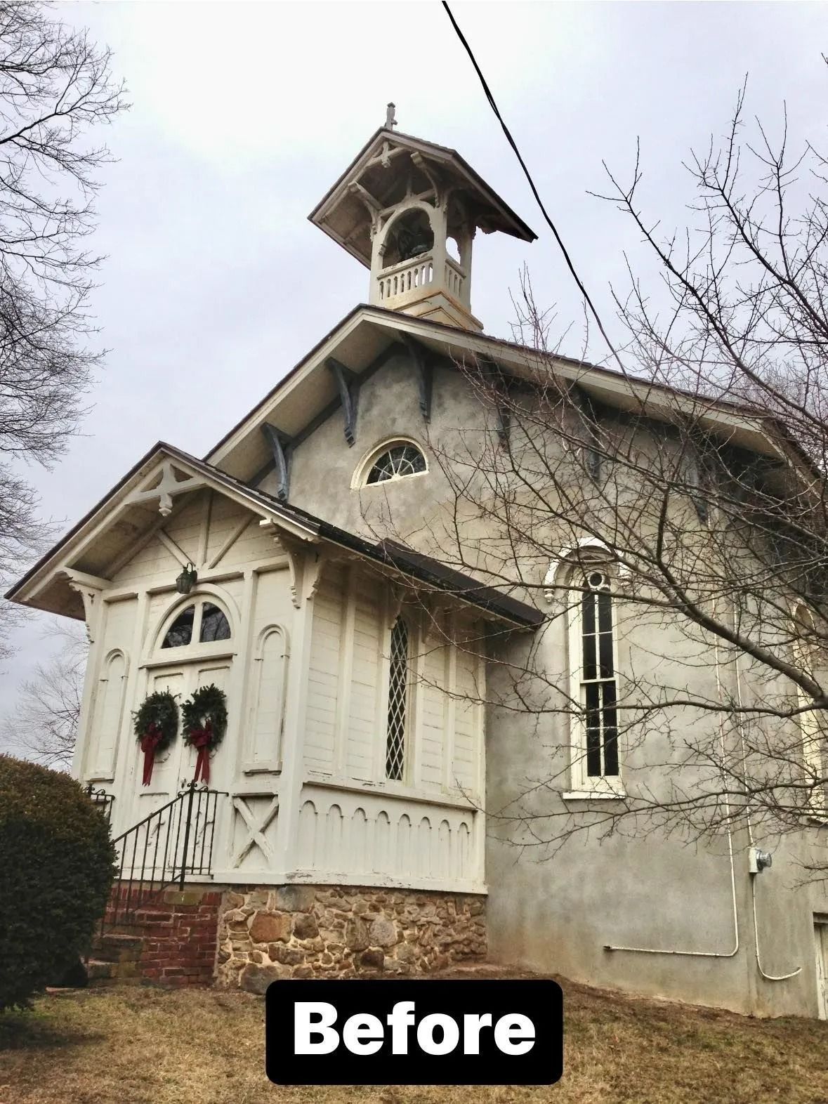 Weathered, light-colored church with bell tower and wreaths over a doorway. The church is in front of bare winter trees.