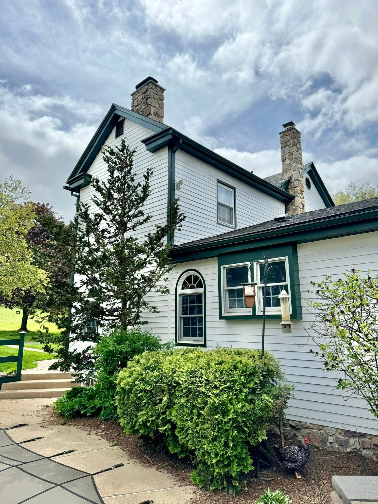 White house with green trim, two chimneys, arched window, shrubbery, cloudy sky.