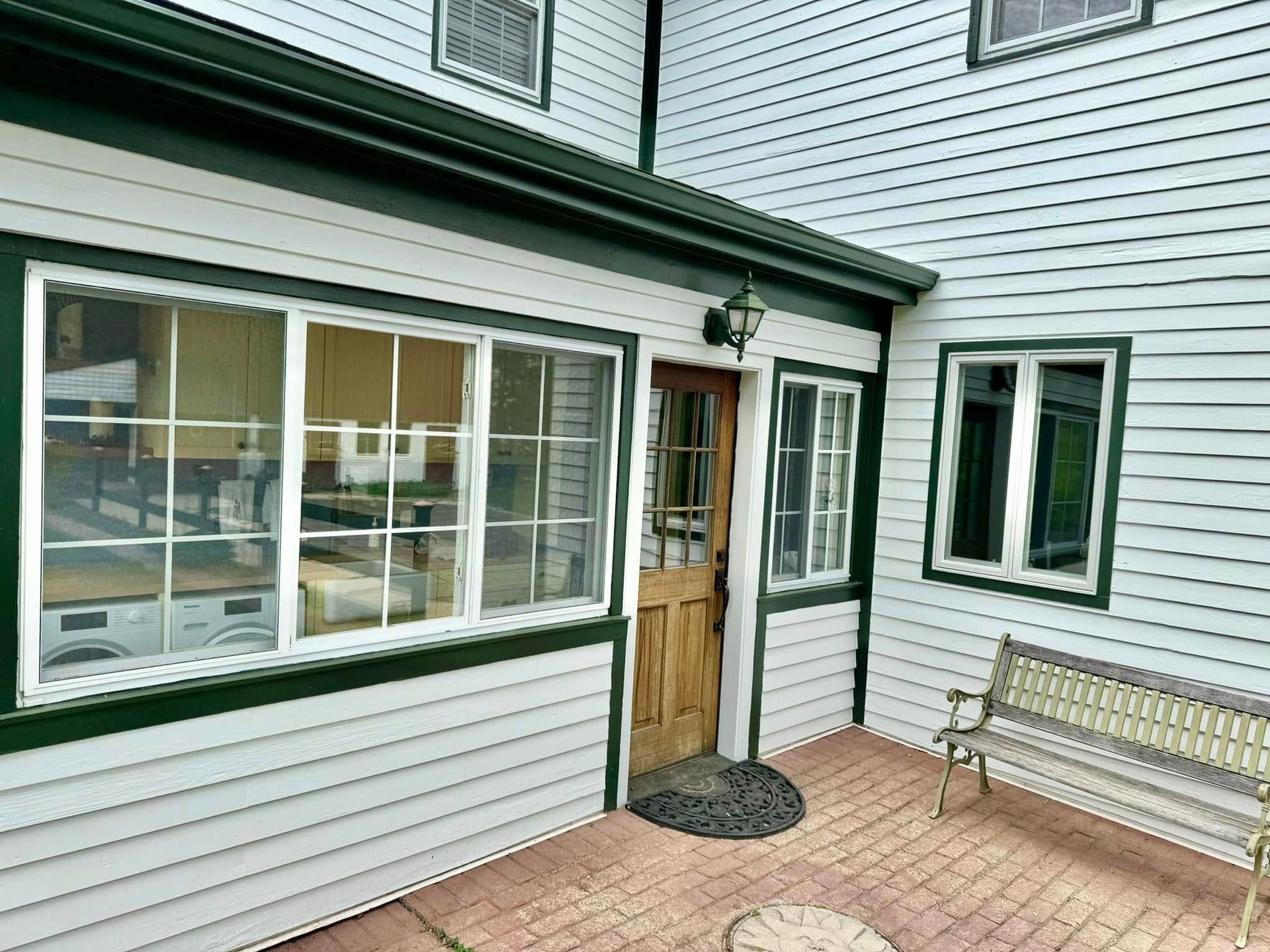 Exterior of a white house with green trim. Windows, a wooden door, and a bench are visible on the brick patio.