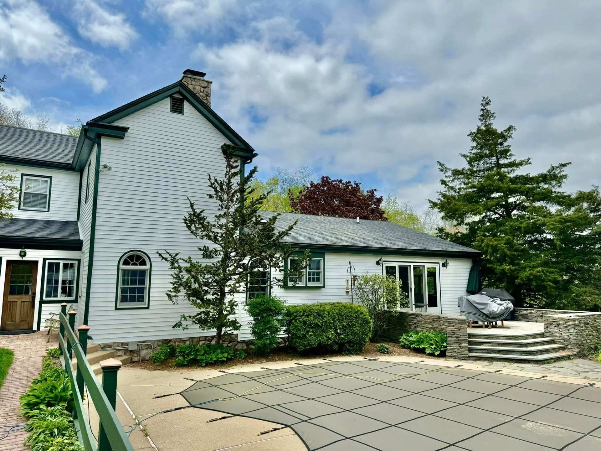 White house with a pool and stone patio. Cloudy sky. Green trim, trees, and pool cover.