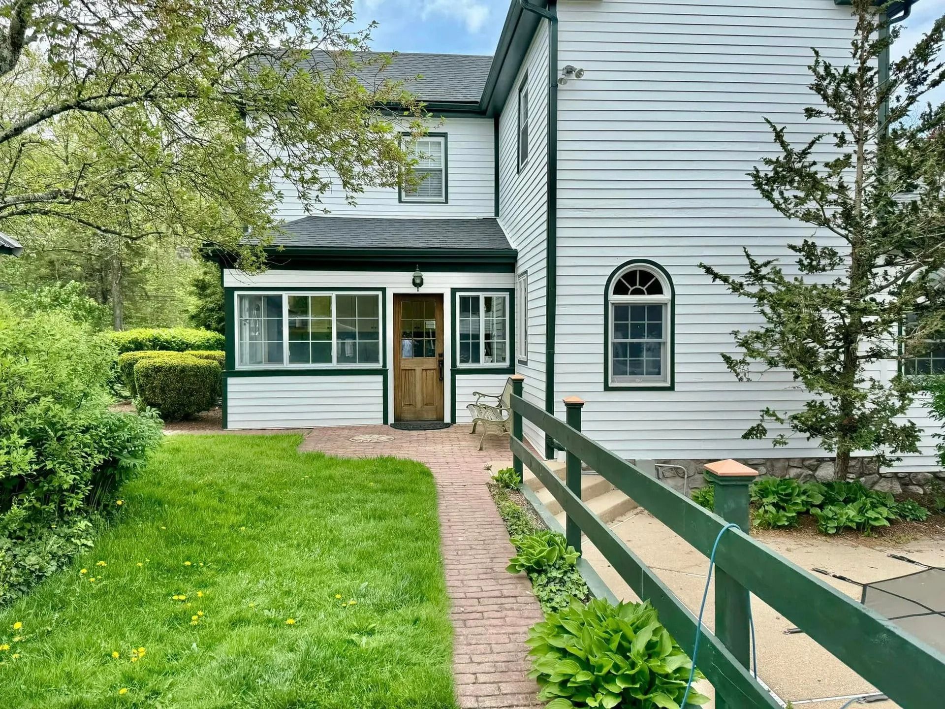 White two-story house with green trim. Brick walkway, green fence, and lawn. Cloudy sky.