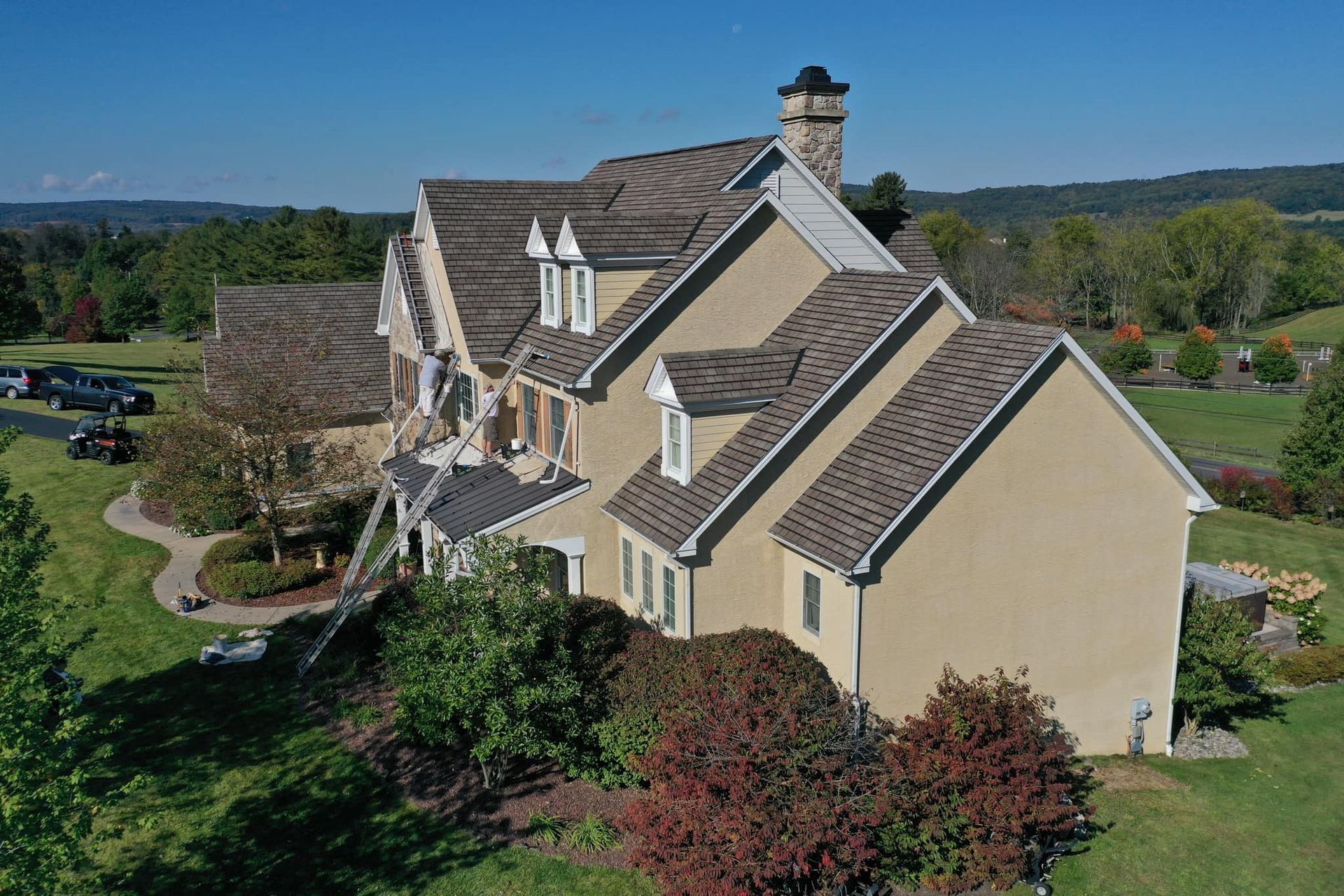 Two-story beige house with brown roof and landscaping. Ladders lean against the roof. Blue sky.