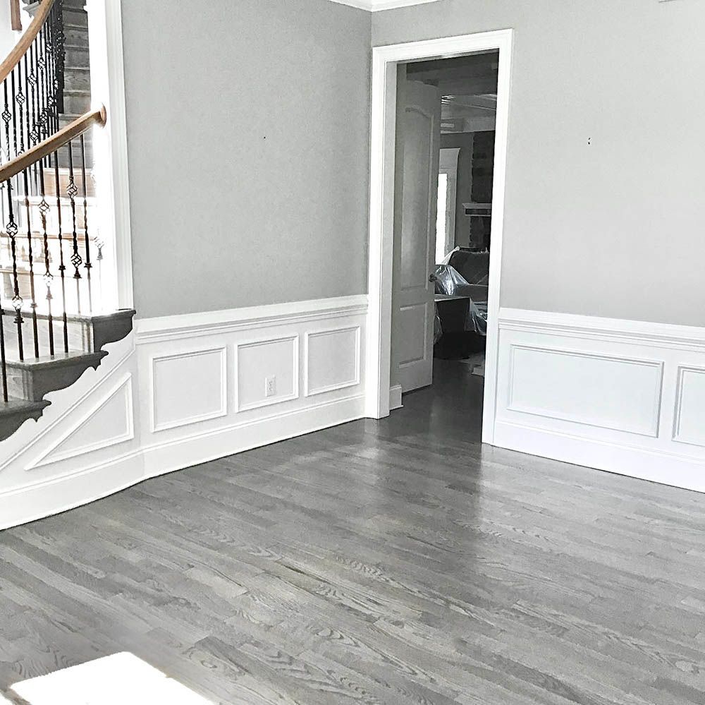 Empty room with gray walls and wood floors, white wainscoting, and a staircase. Doorway leads to a cluttered area.