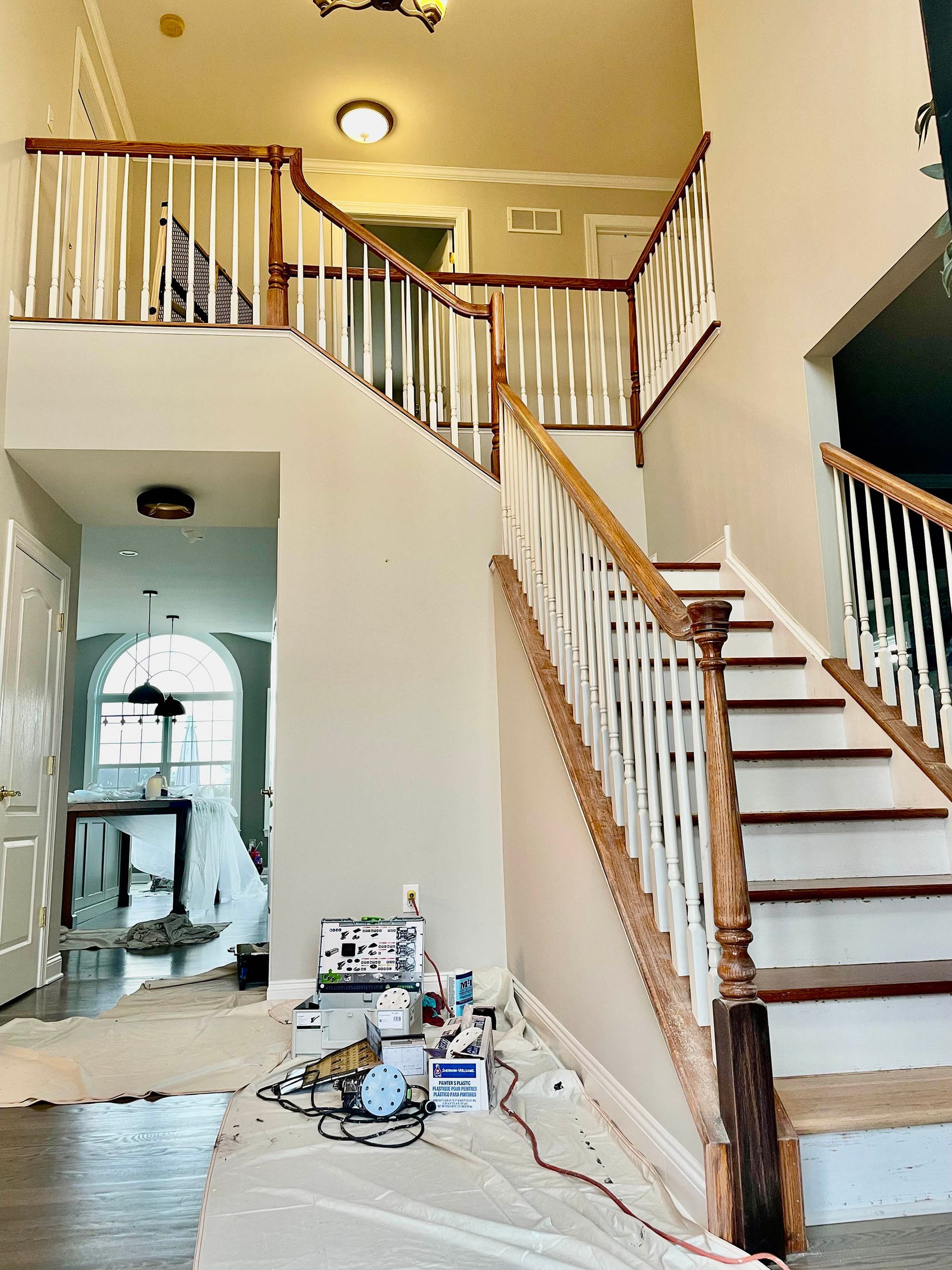 Two-story foyer with staircase. White spindles, wood handrails, neutral walls. Painting supplies on floor.