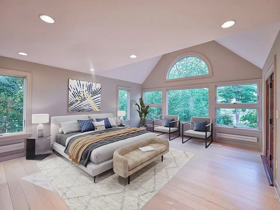 Bedroom with a bed, bench, and two chairs. Large windows offer a view of trees.