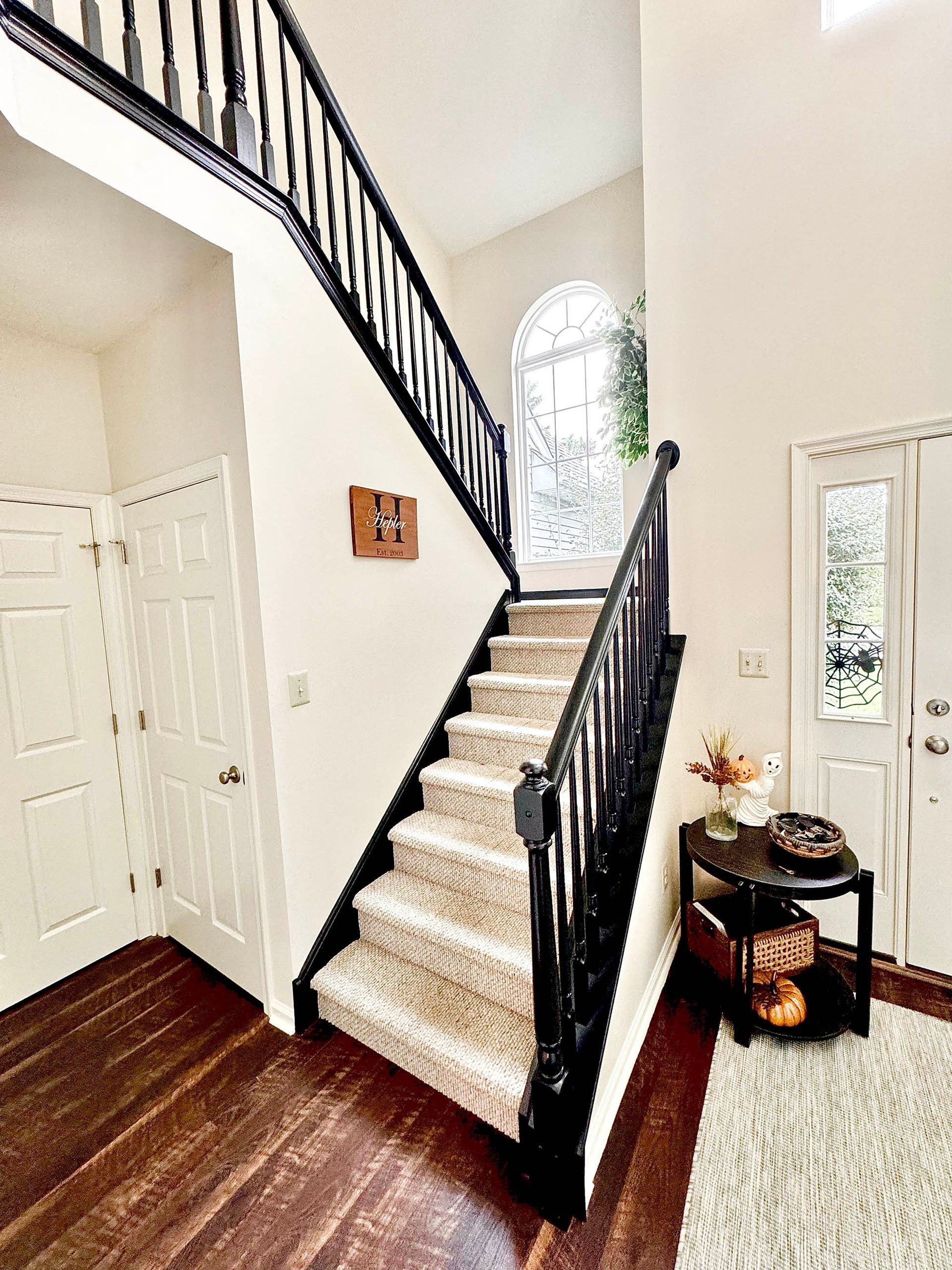 Staircase with black banister, light carpet steps, and dark wood floors. A small table with décor is near the entry door.