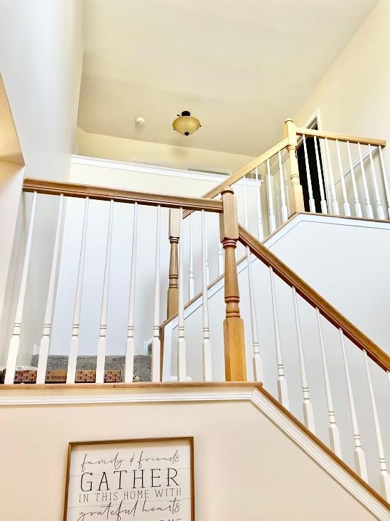 Staircase with wooden handrails and white balusters. Light fixture on high ceiling. Framed sign at the bottom.