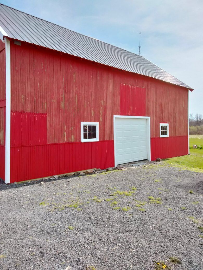 Red barn with a white garage door, small windows, and a gravel driveway under a blue sky.