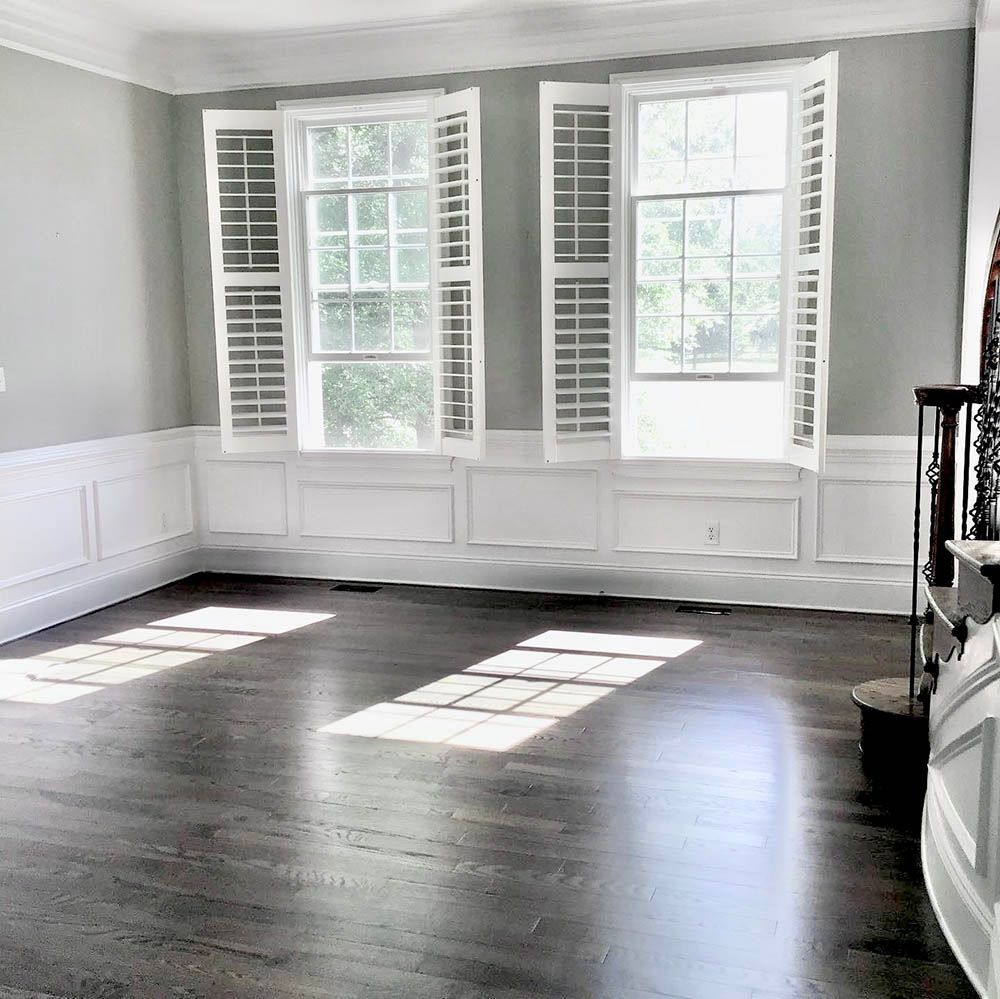 Empty room with hardwood floors, gray walls, white trim, and two windows with shutters. Sunlight streams in.
