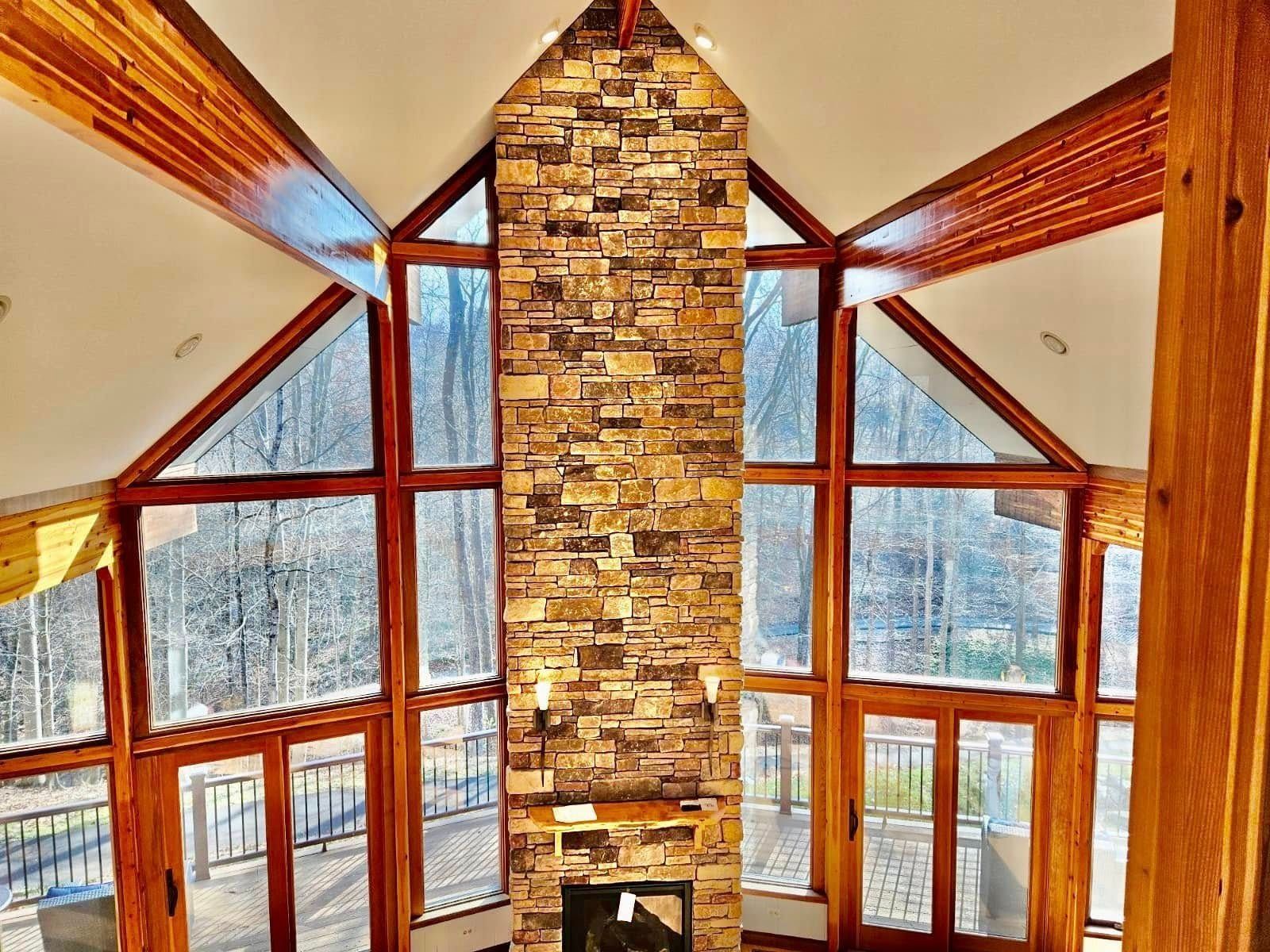 Stone fireplace with large windows in a cabin, wooden beams, and a view of trees.