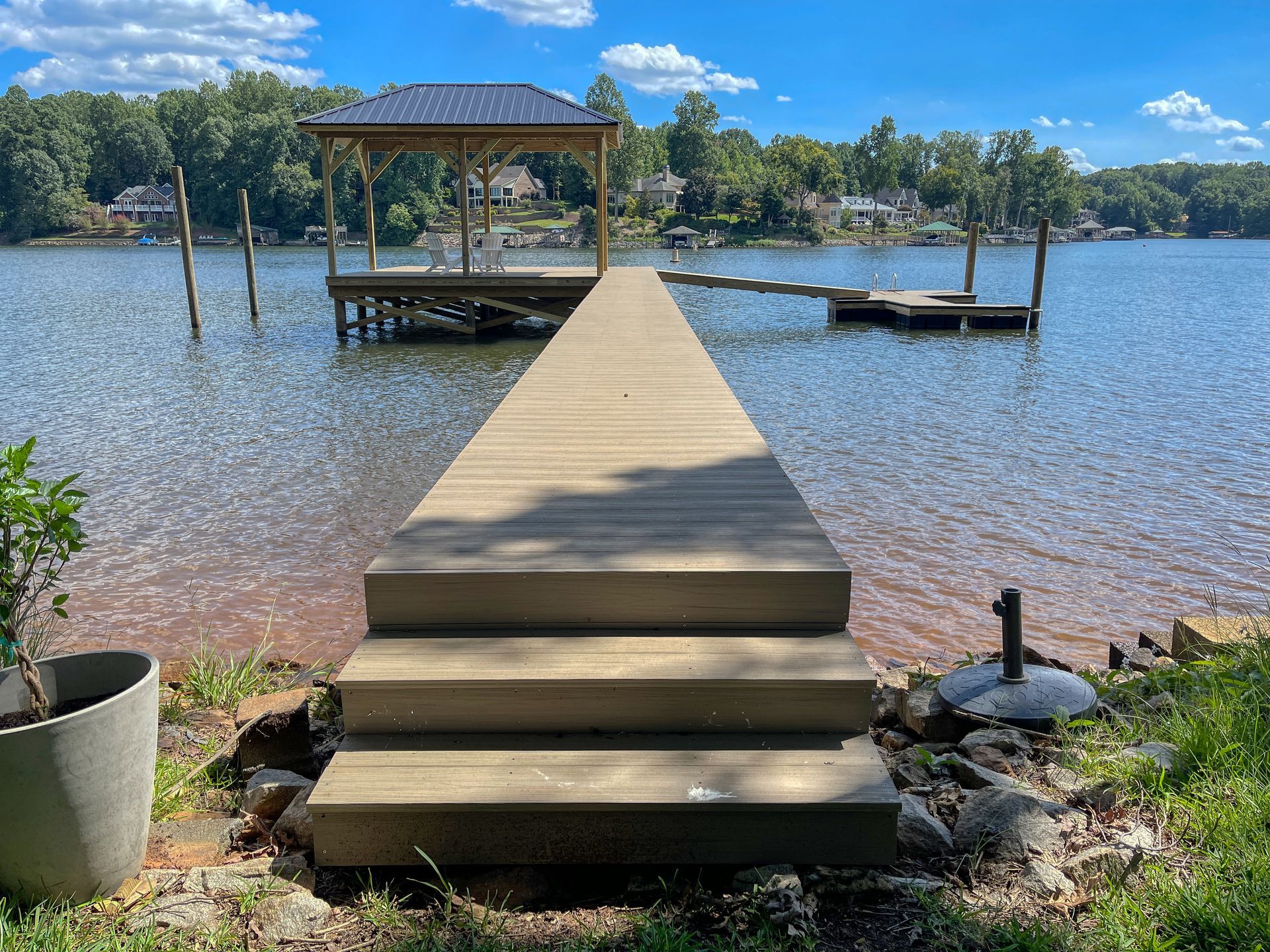 Wooden dock with stairs leading to a gazebo over calm water, surrounded by trees under a blue sky.