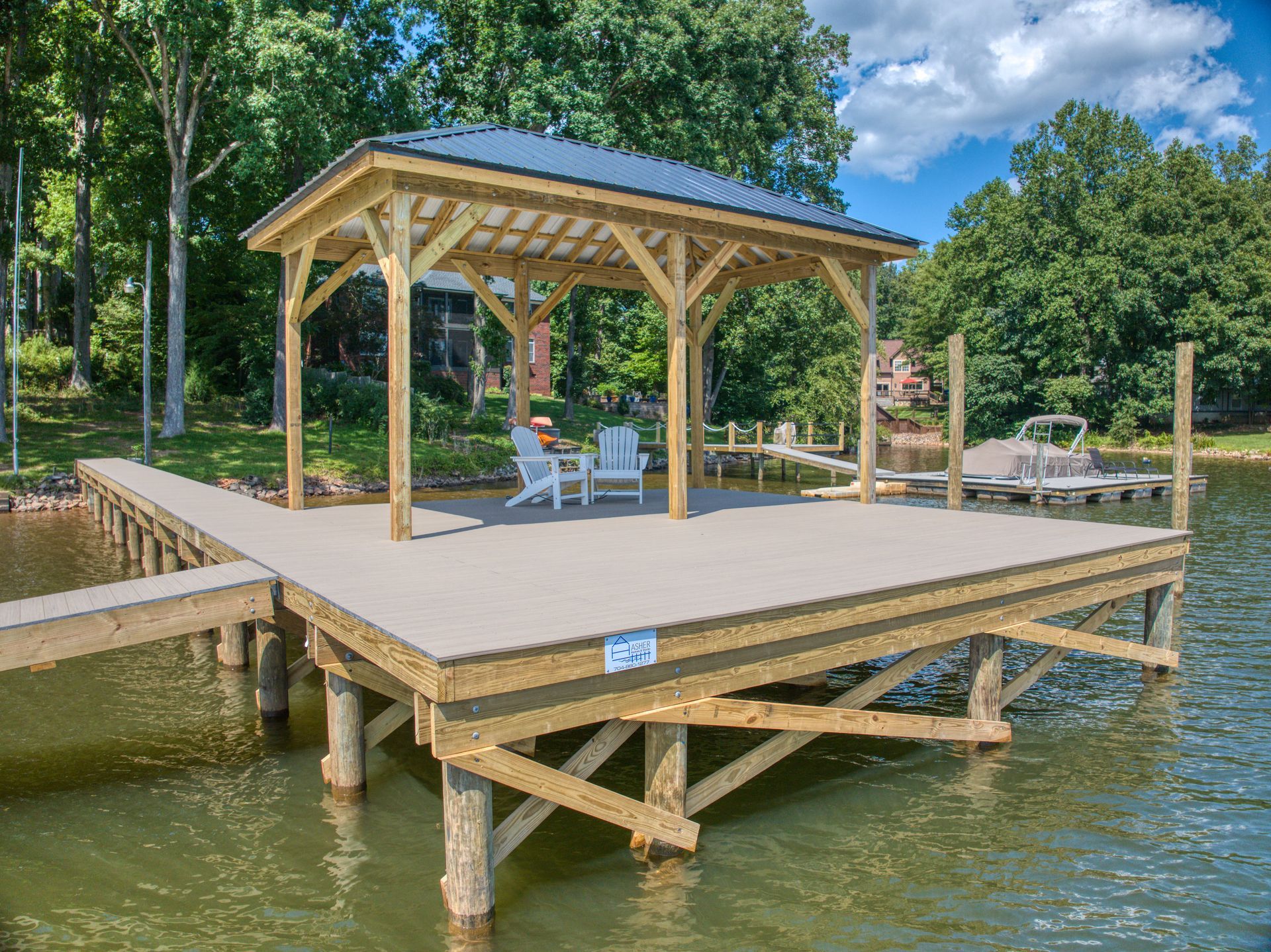 Wooden dock with gazebo on a lake. White chairs and table under the gazebo. Green trees and blue sky.