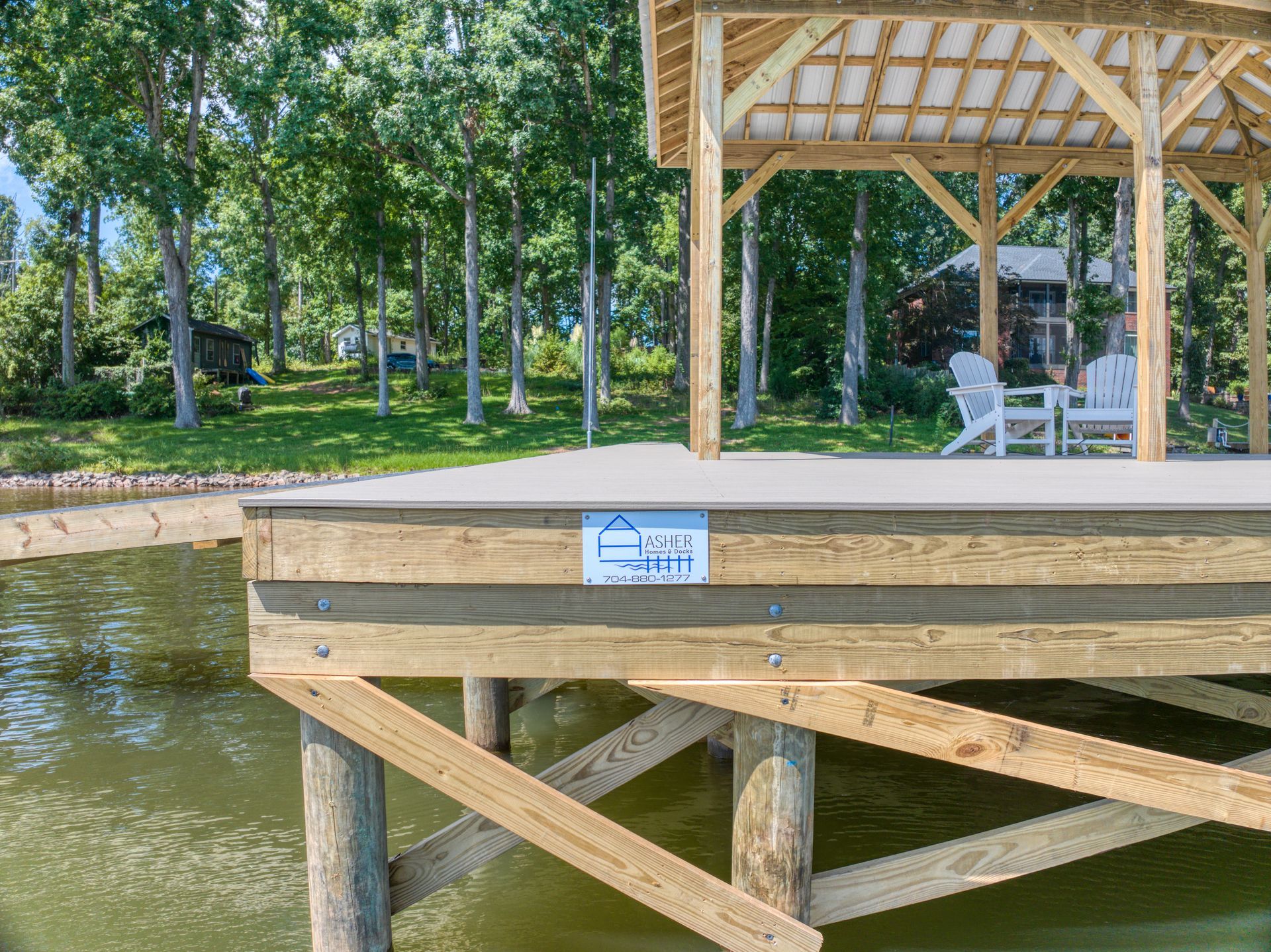 Wooden dock with gazebo, overlooking a lake. Two white chairs under gazebo. Green trees in background.