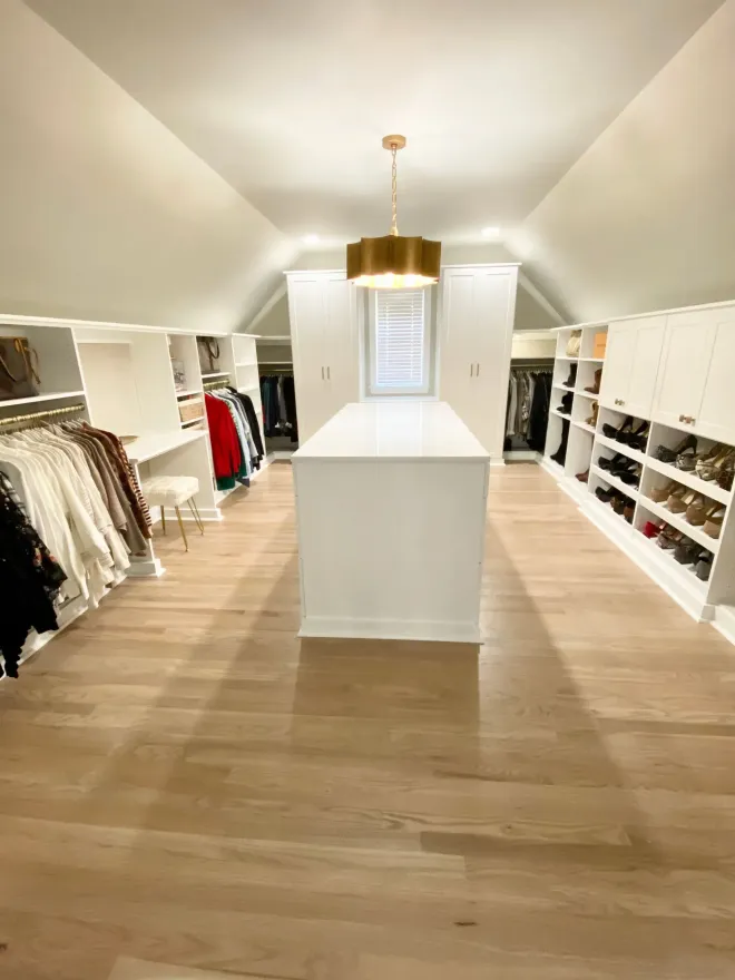 Spacious, white-walled walk-in closet with island, clothing racks, shoe shelves, and a gold pendant light.