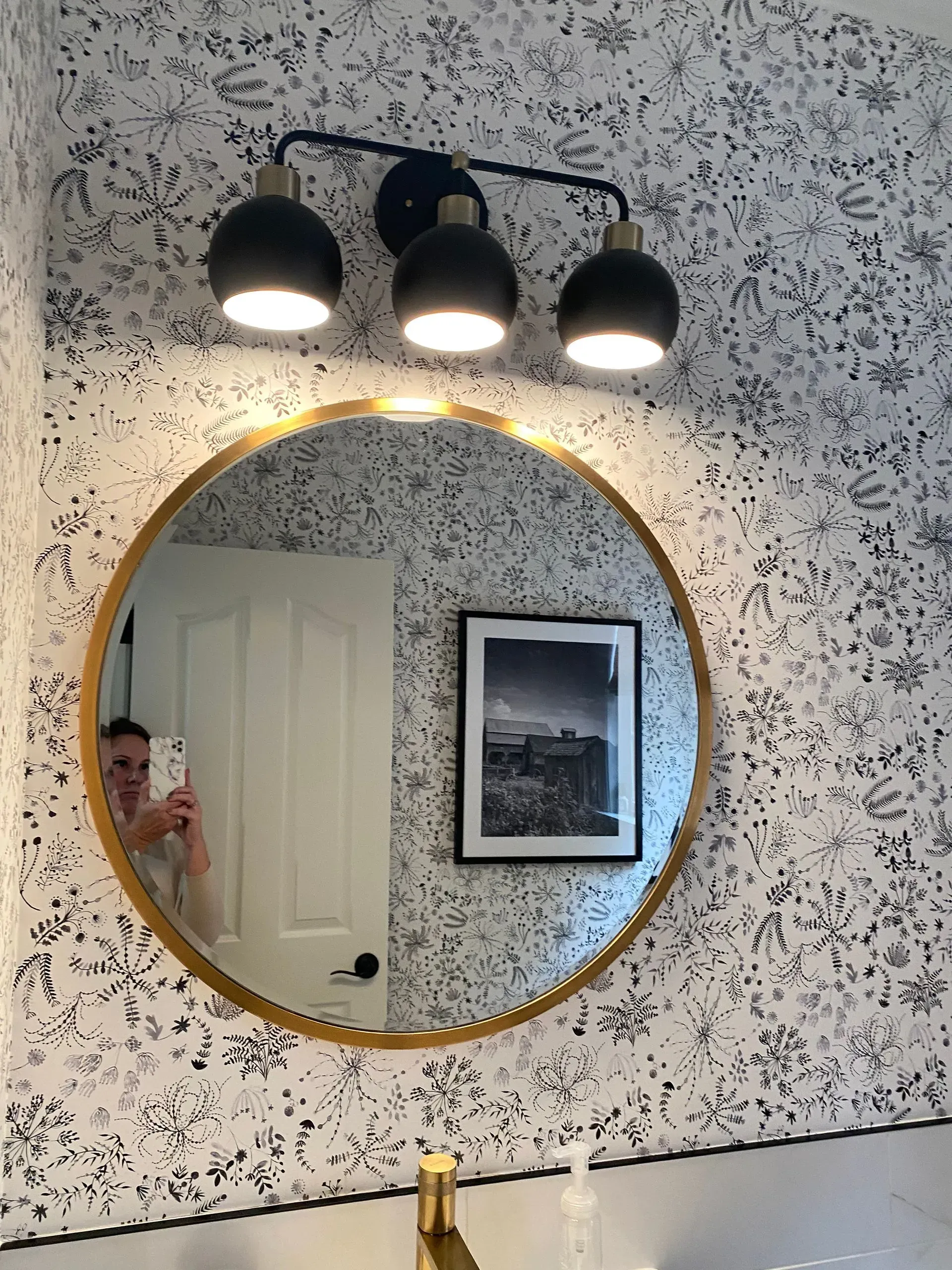 Bathroom with round gold mirror, black and gold light fixture, patterned wallpaper, and framed photo.