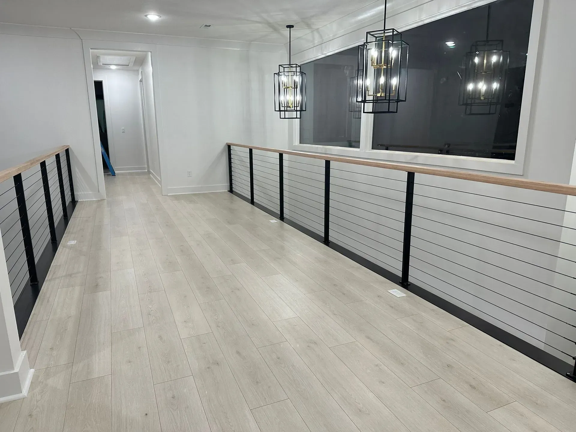Hallway with light wood floors, white walls, black railings, and hanging lights.