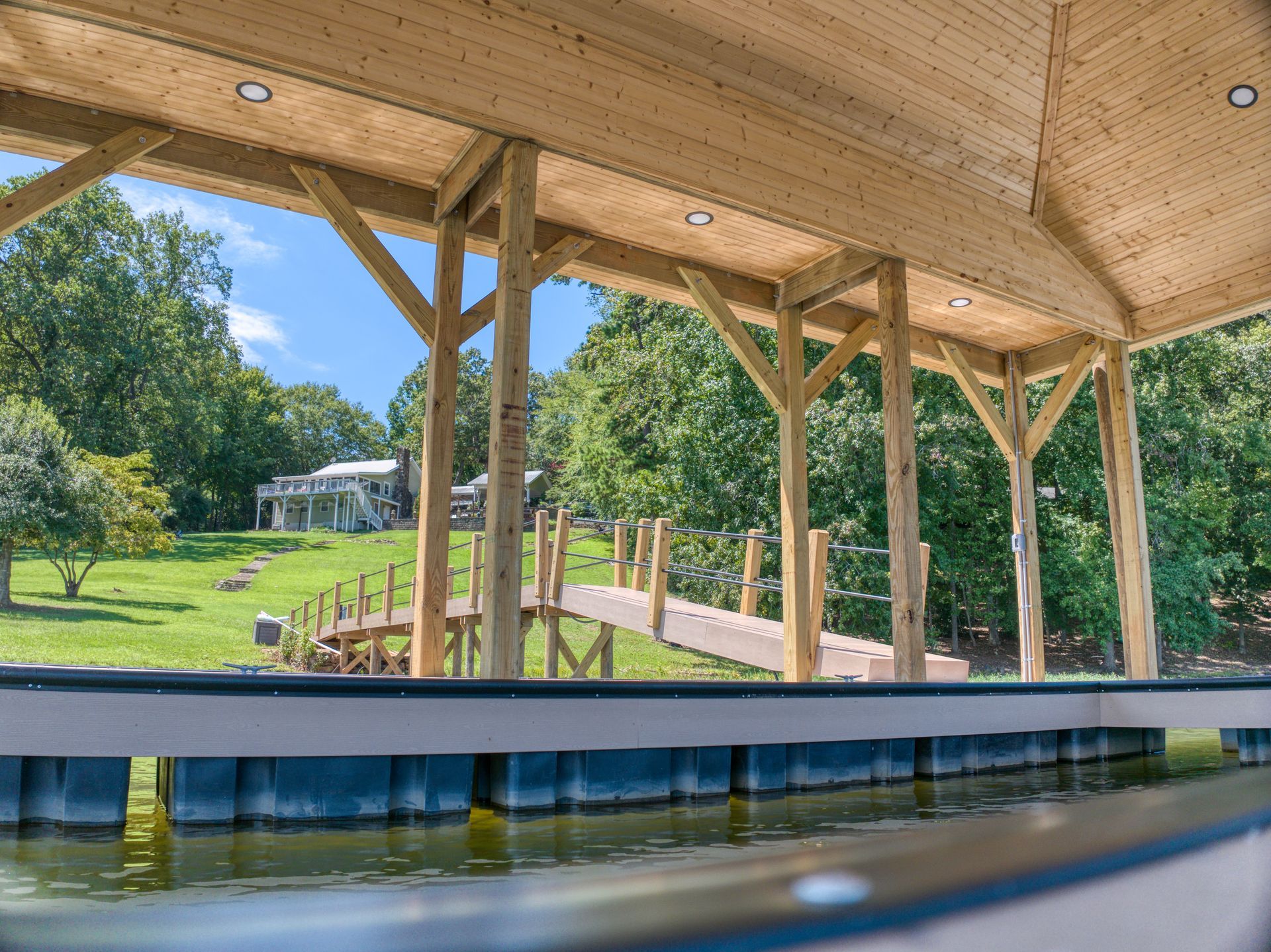 Wooden boat dock structure with a pier leading to a house on a grassy hill, under a clear sky.