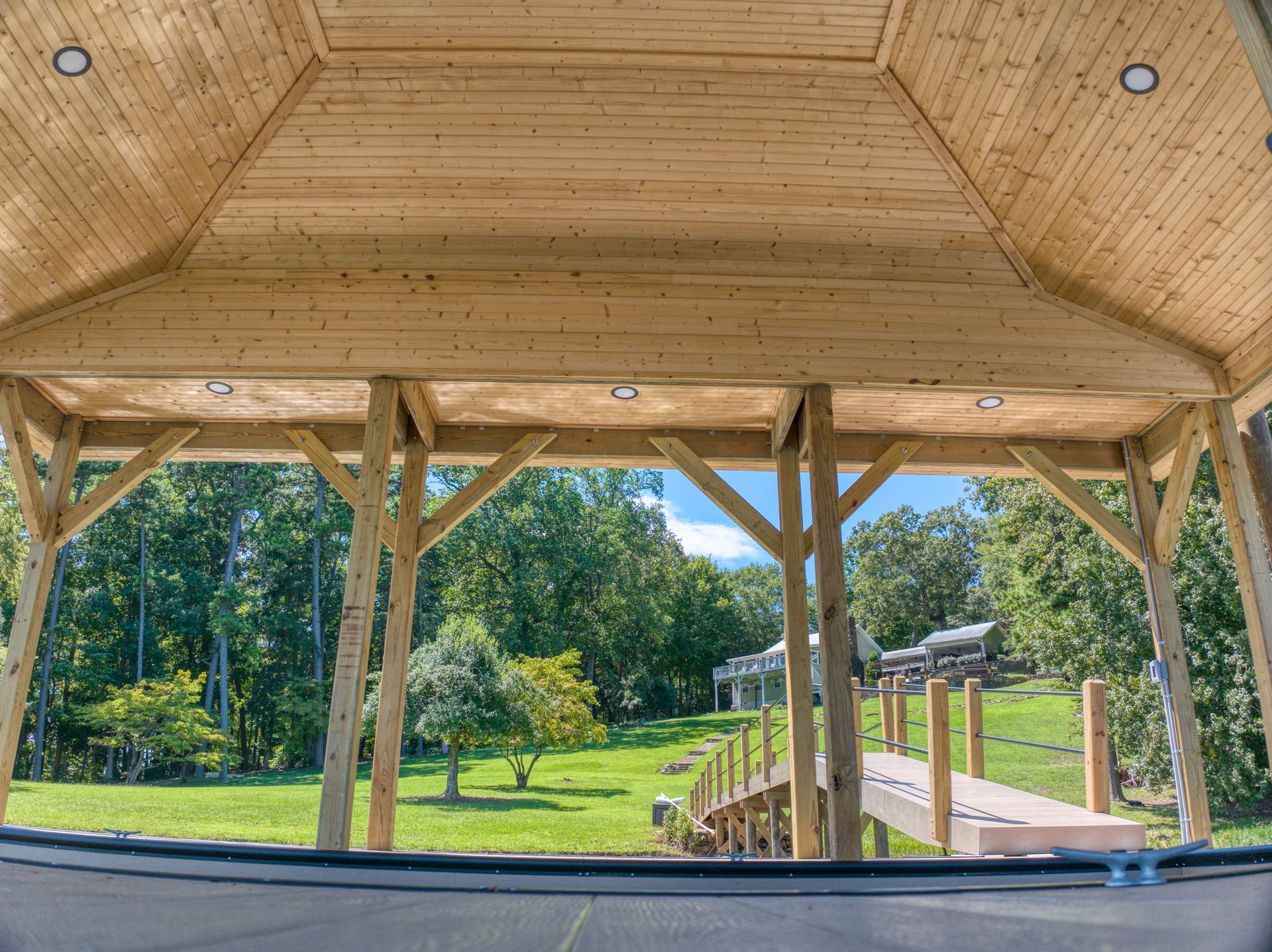 Wooden gazebo overlooking a grassy lawn, trees, and a wooden dock on a sunny day.