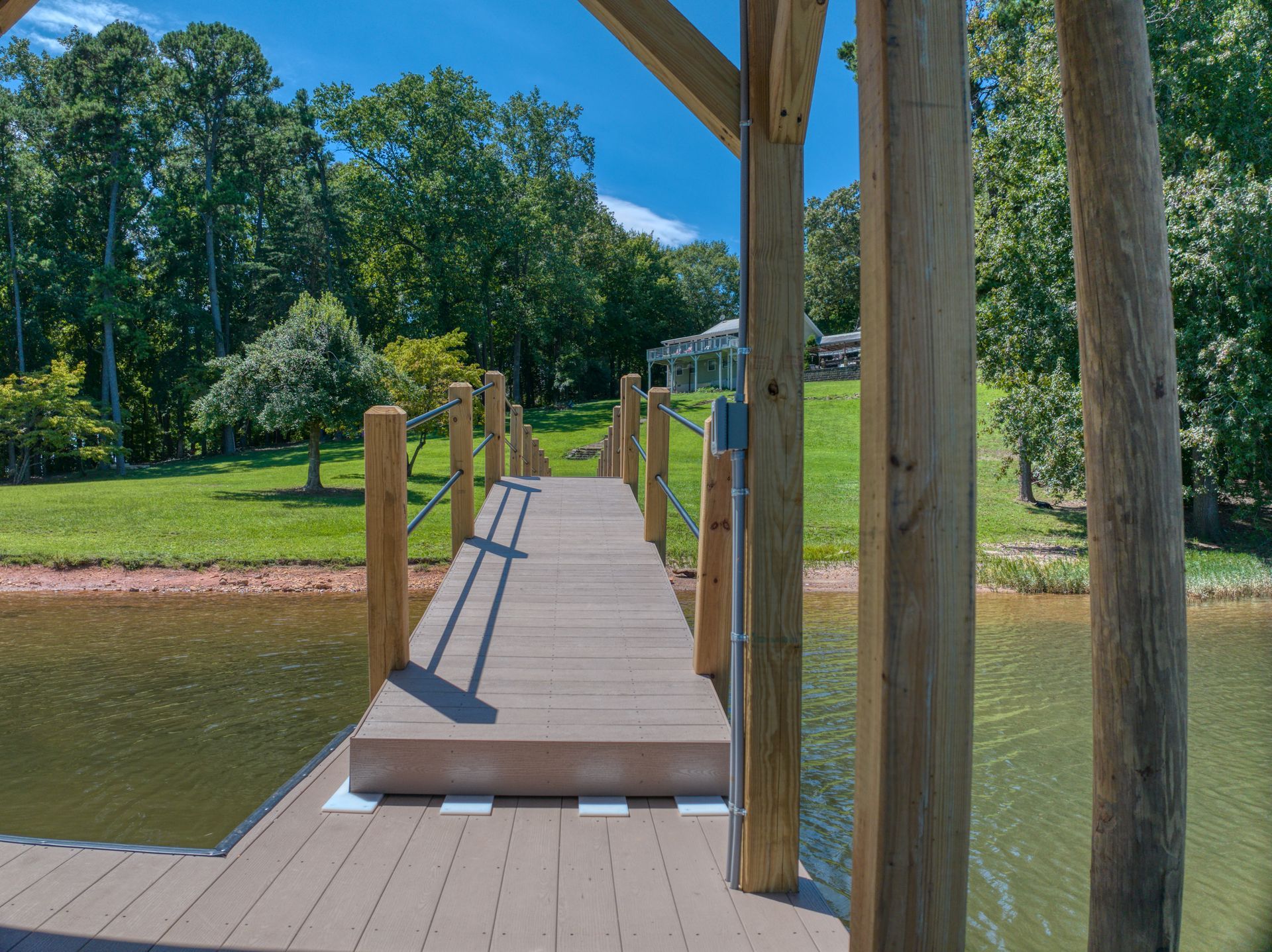 Wooden dock extending over water toward a grassy bank with trees and a house in the distance.