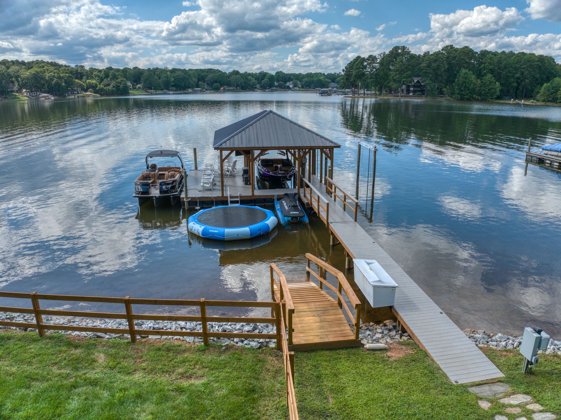 Dock with boat, boat house, and floating trampoline on a lake under a cloudy sky.