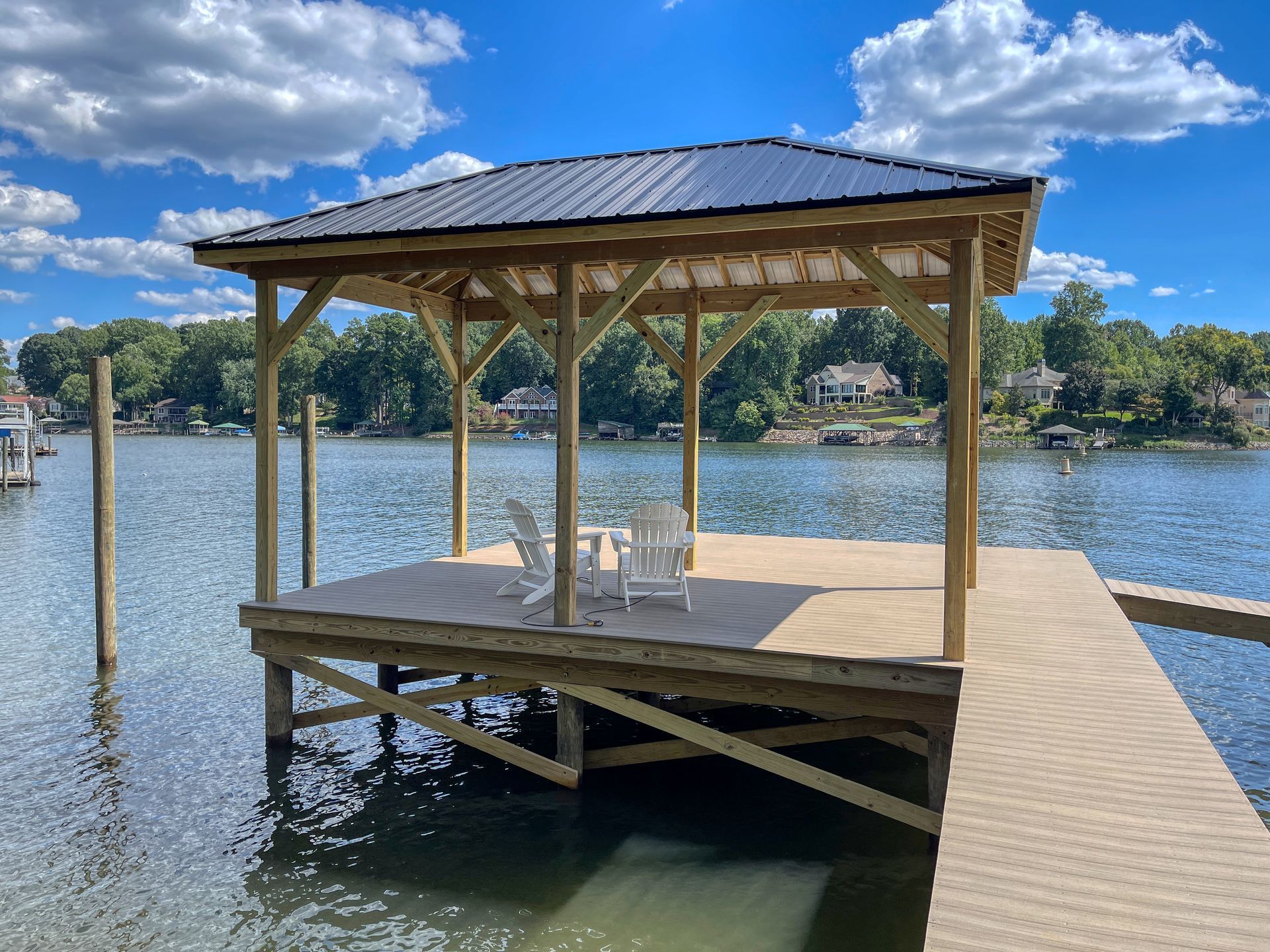 Wooden gazebo with metal roof on a dock over water with two white chairs.
