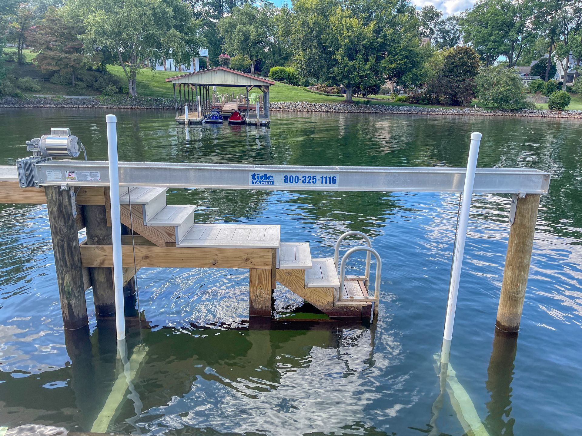 A wooden dock with steps leading into water, with a boat lift and a boathouse in the distance.
