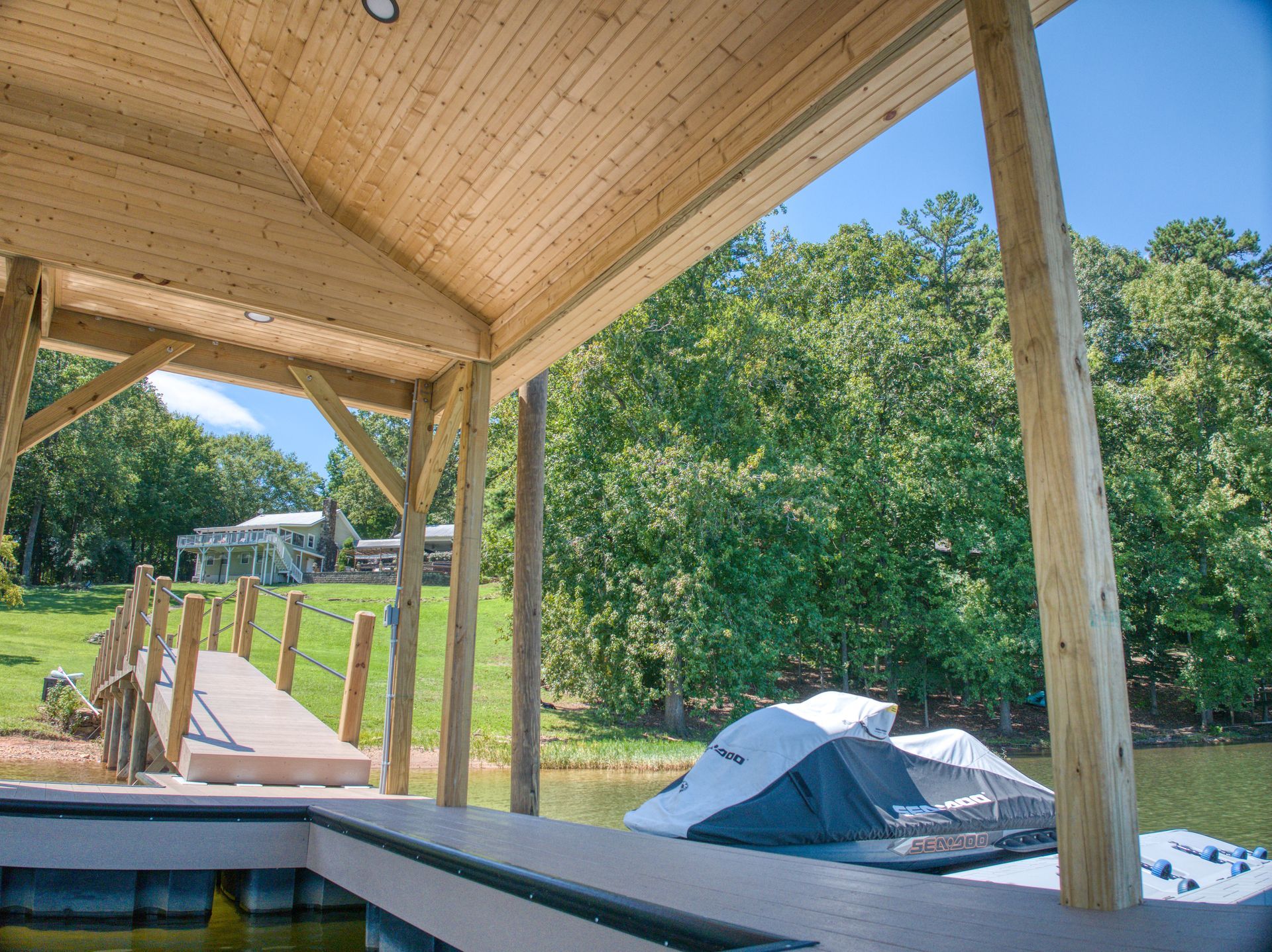 Wooden dock with covered area and boat, overlooking green trees and a house.