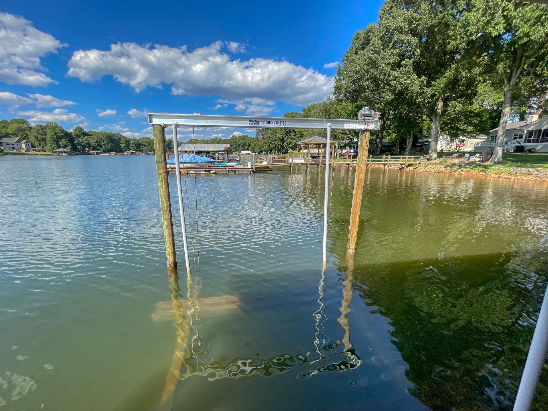 A boat lift structure partially submerged in a lake. Bright blue sky with clouds, trees and homes in the background.