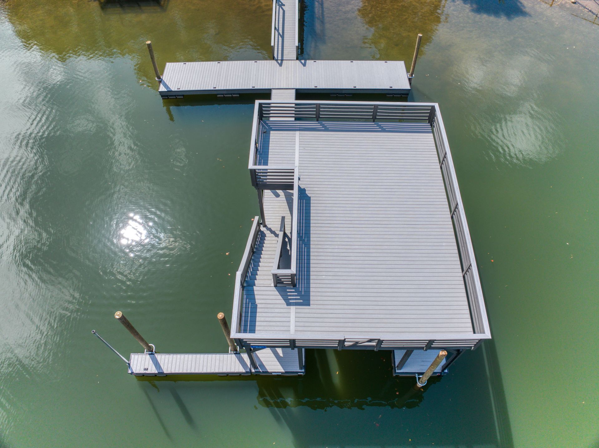 Overhead view of a gray wooden dock complex on green water. Sun reflects off the water's surface.