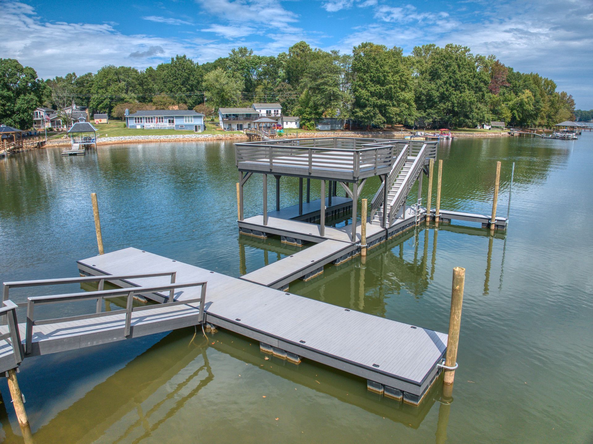 Dock extending into a lake, with stairs leading up to a covered platform, houses in the background.