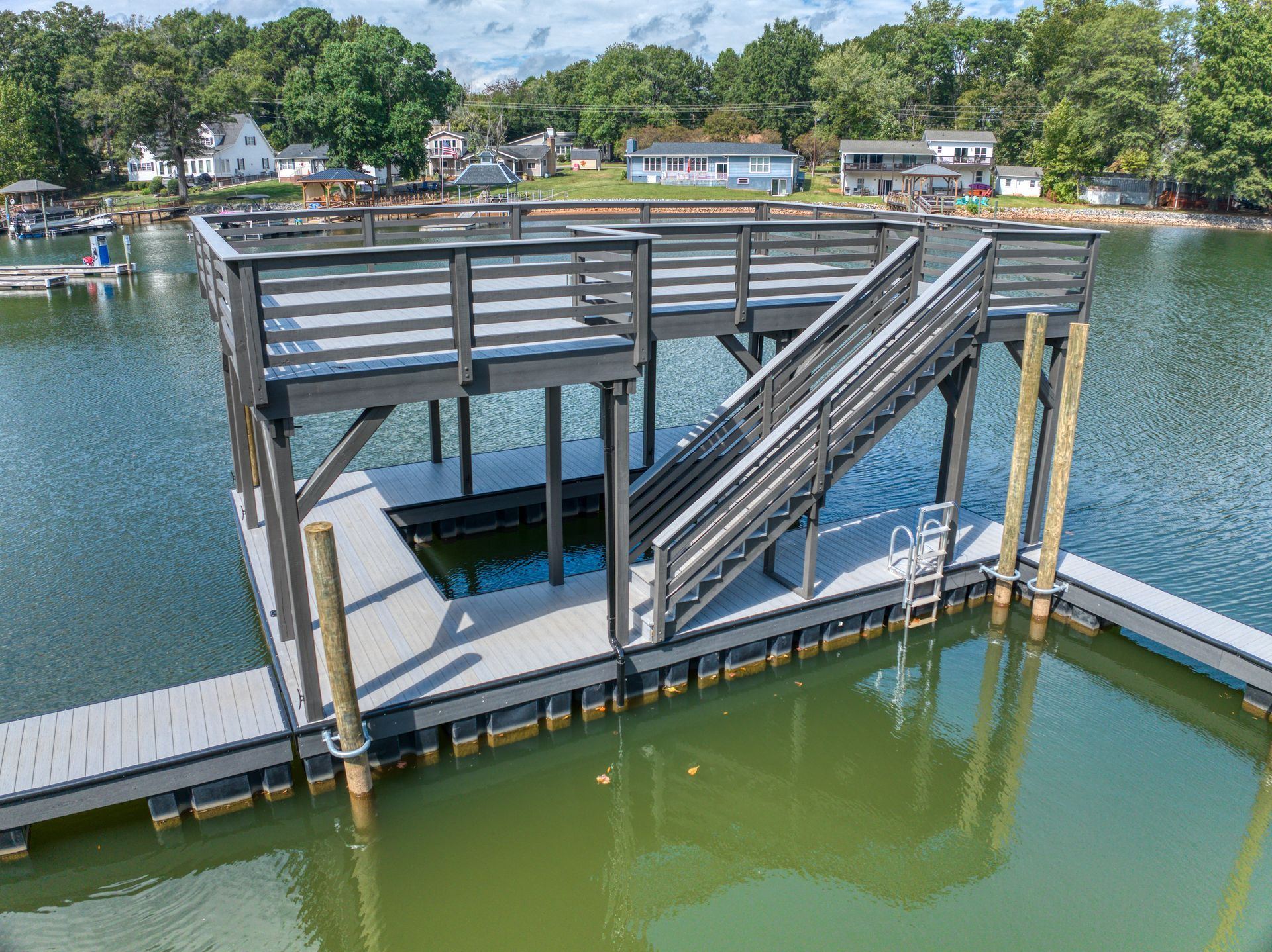 Dock with stairs and elevated platform on a lake; gray and brown tones; surrounding houses and trees.