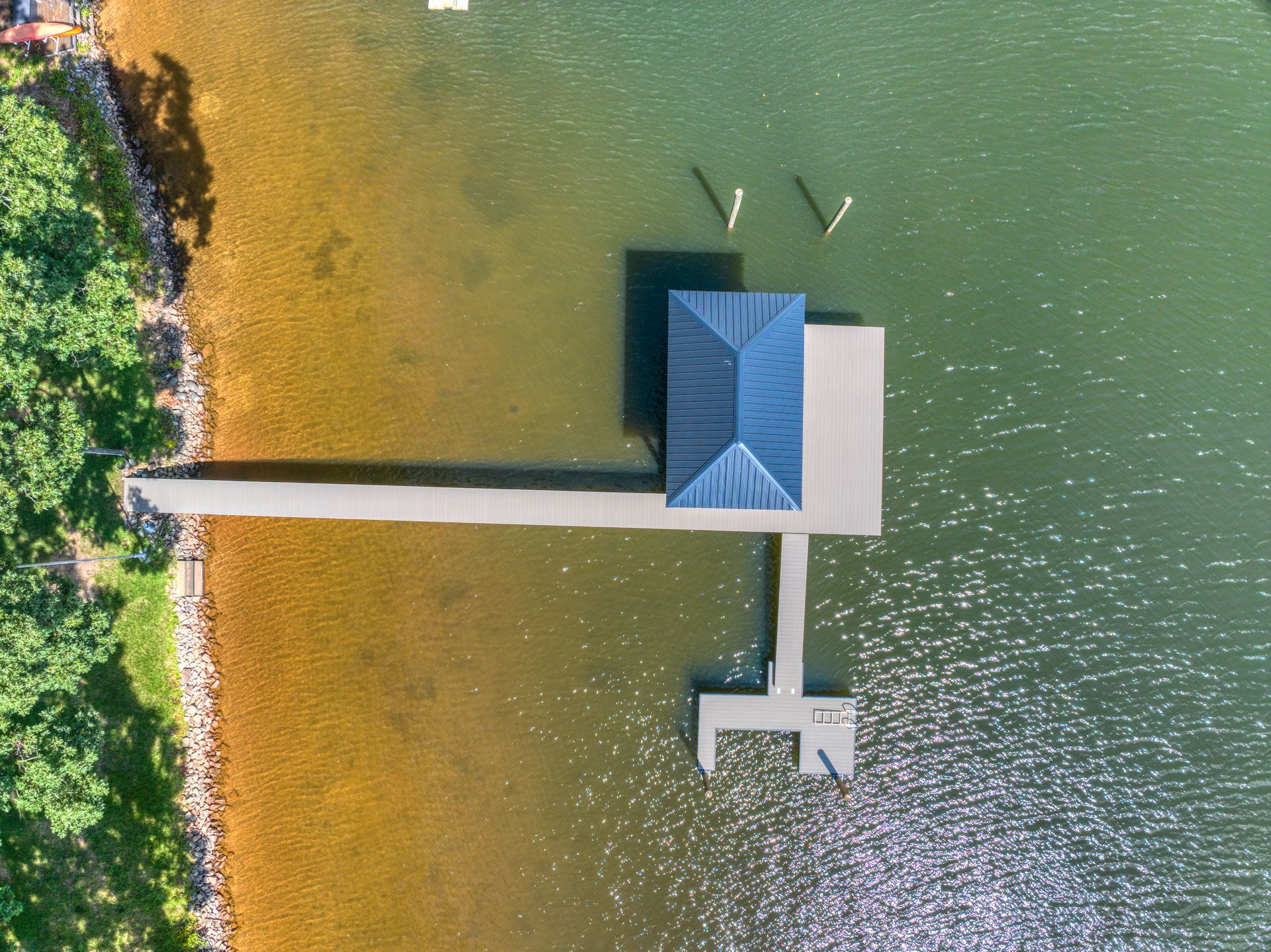 Overhead view of a dock with a blue-roofed shelter extending into a lake. The water transitions from sandy to green.