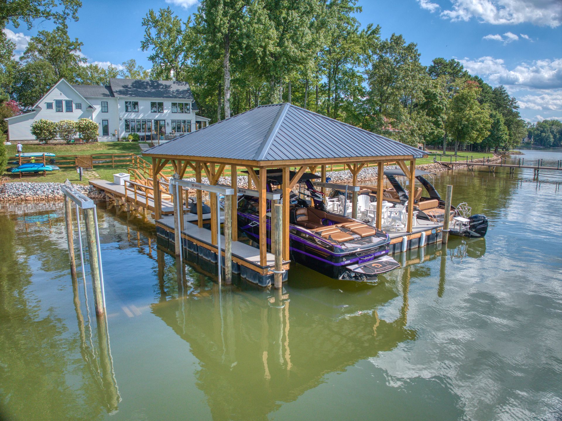 Dock with boat under a gray metal roof, overlooking lake with large house in the background.