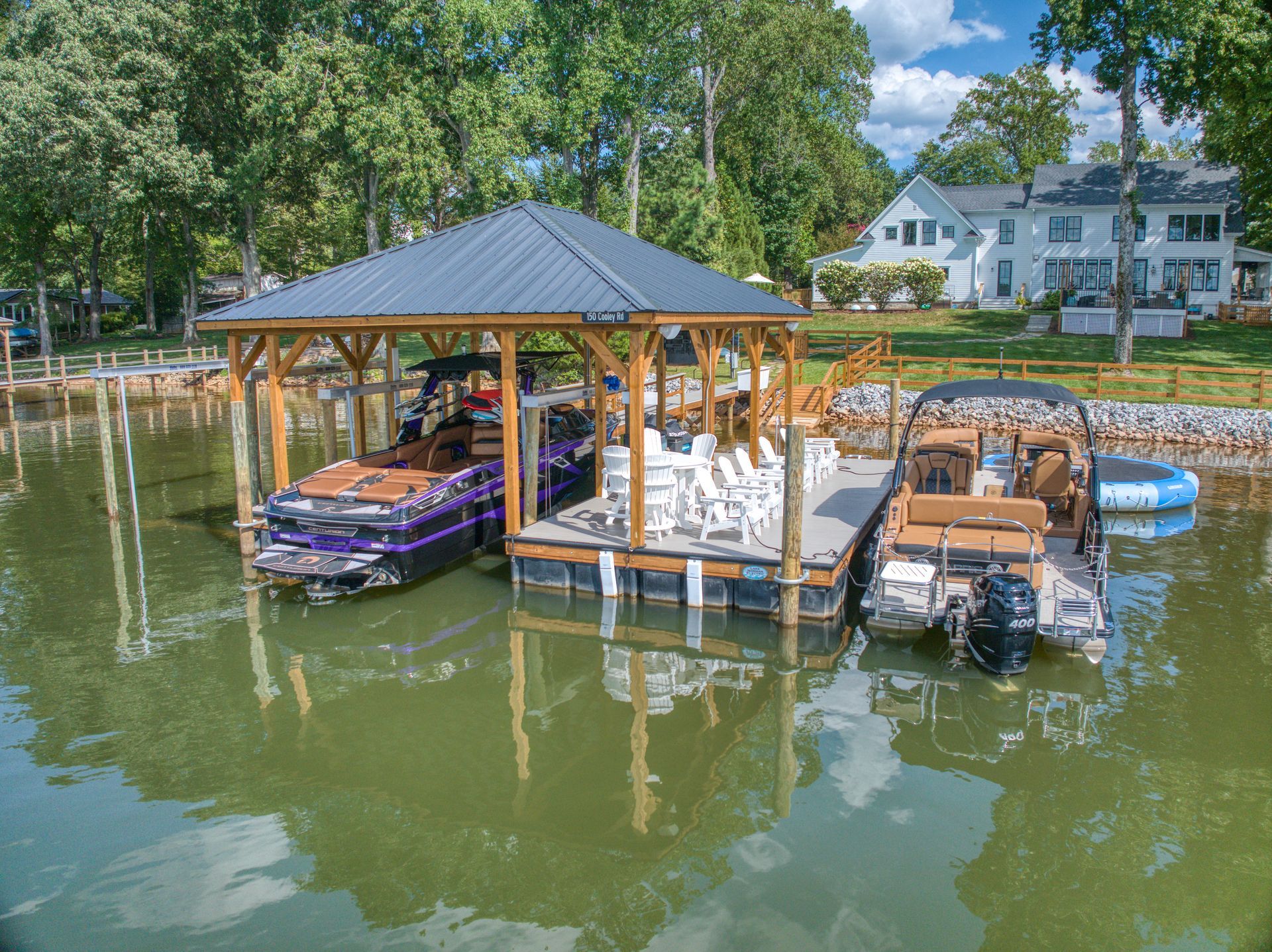 Boats docked at a wooden pier with a covered shelter, on a lake near homes under a blue sky.