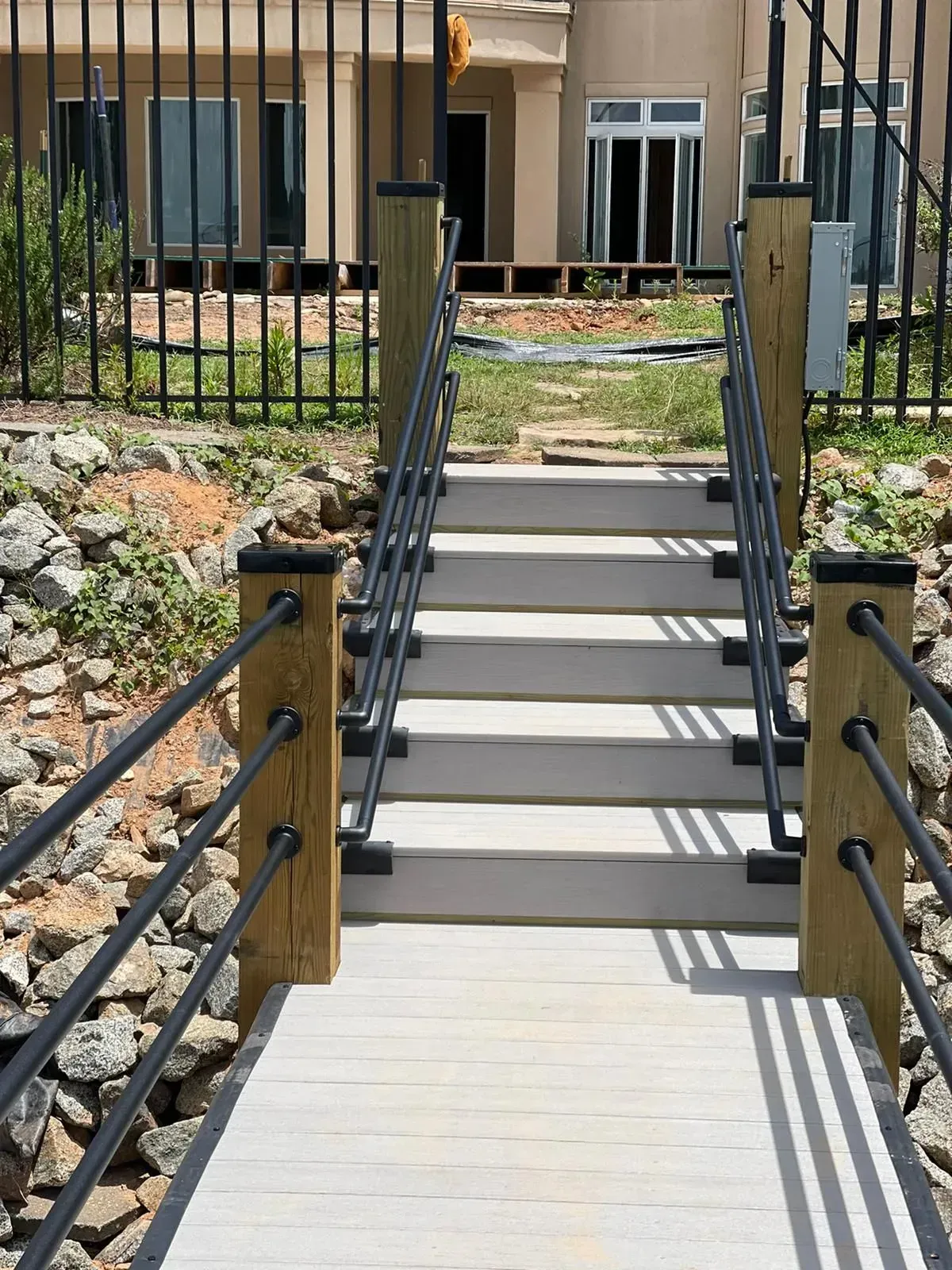 Wooden bridge with black railing, steps lead up to a building in the background.