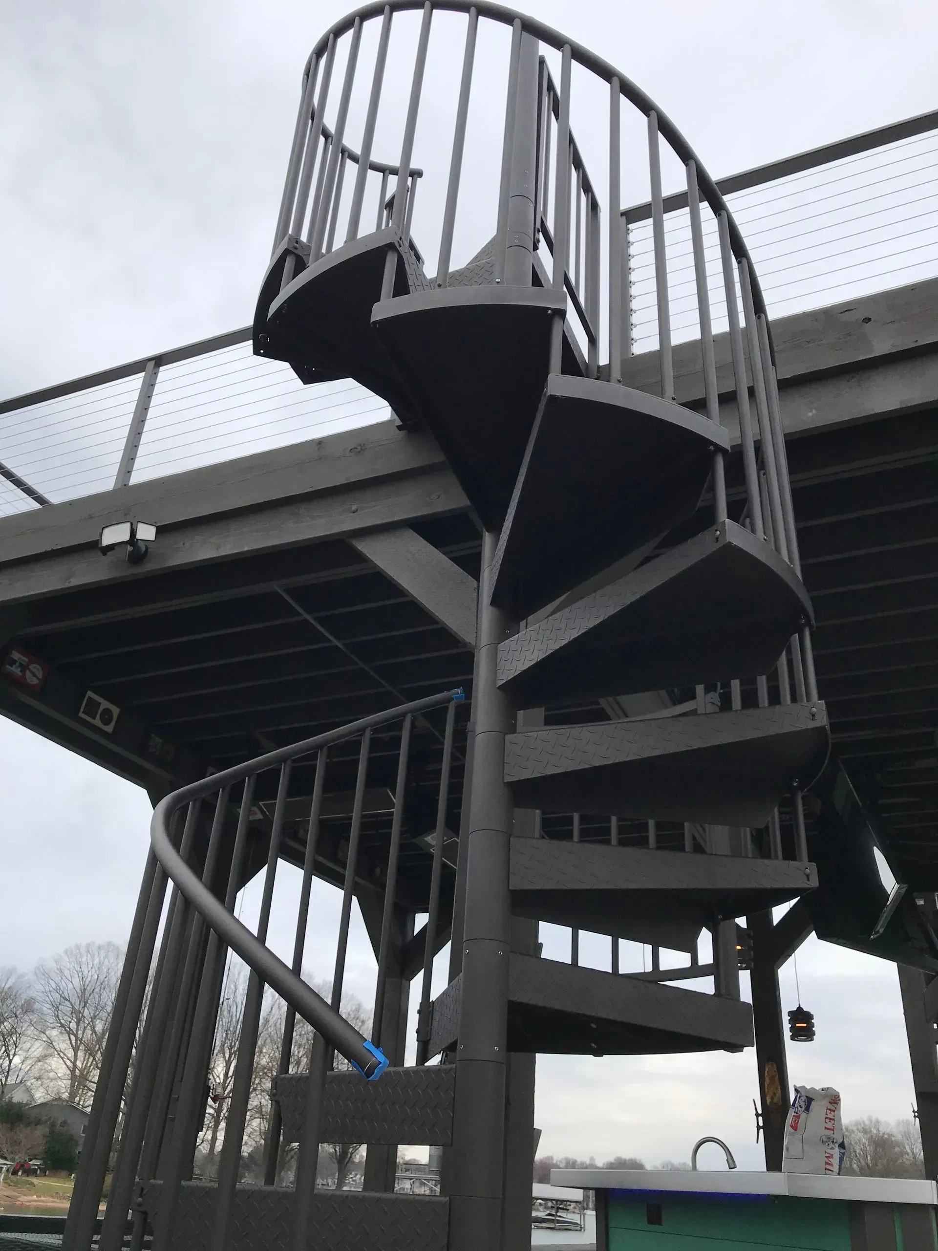 Dark grey spiral staircase leads to an elevated wooden platform; overcast sky in the background.