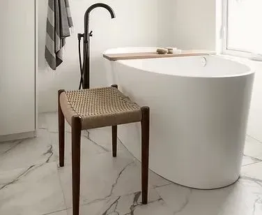 Bathroom with white oval tub, woven stool, dark faucet, and marble floors.