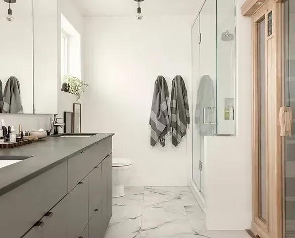 Modern bathroom with gray vanity, white walls, glass shower, and marble floors. Two towels hang.
